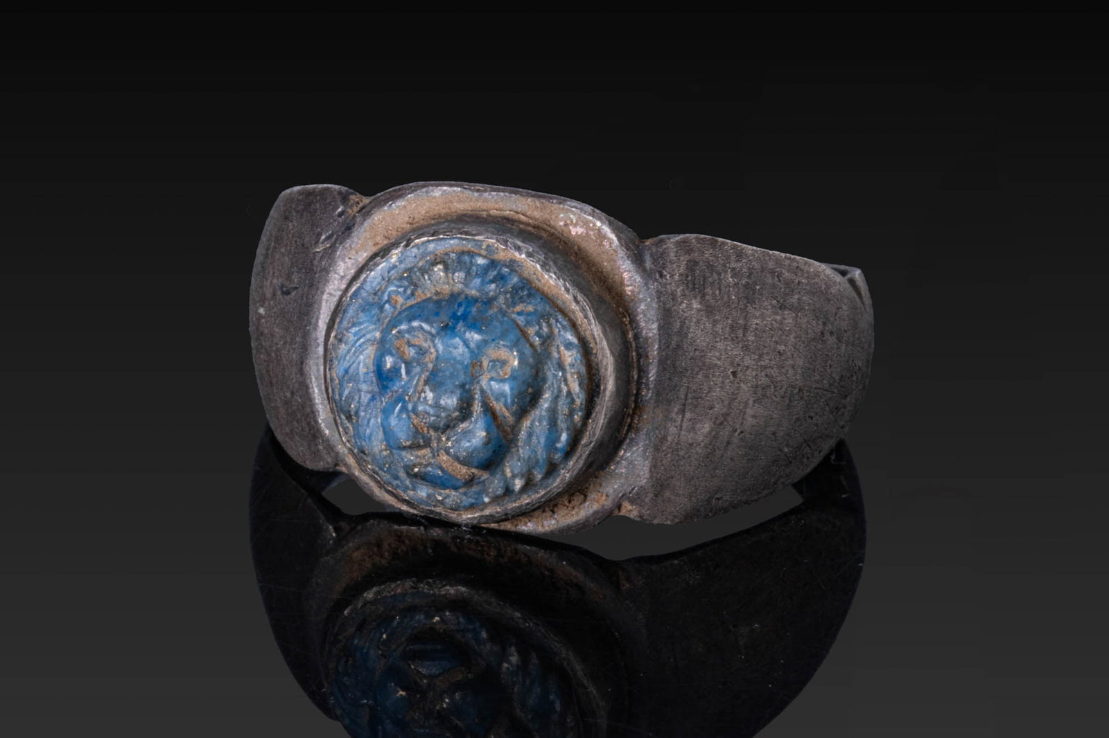 ROMAN SILVER RING WITH GLASS CAMEO OF A LION?S HEAD: Ca. AD 300. A silver ring with a rounded hoop, expanding shoulders and a flat bezel plate with a circular mount set with a circular glass cameo carved as a lion's head with a prominent muzzle, whisker