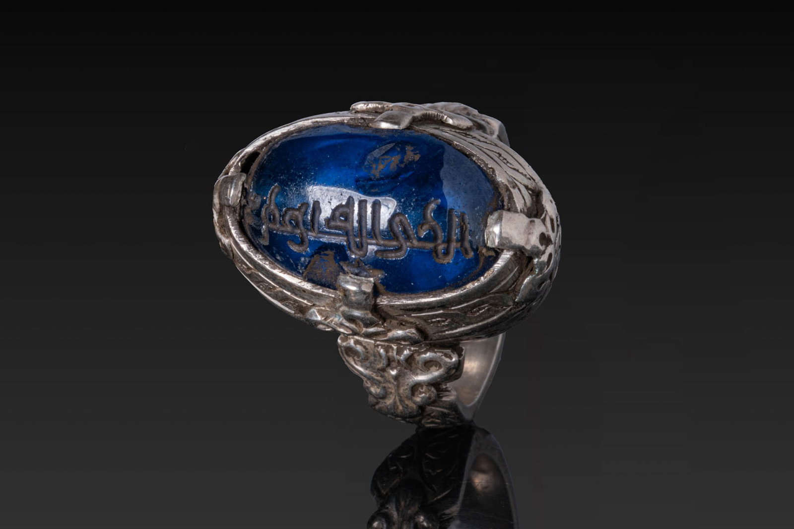 SASANIAN INSCRIBED BLUE INTAGLIO IN SILVER RING: Ca. AD 300 - 600. A cobalt blue oval intaglio engraved with a line of script, set in a silver ring with a high oval collet framed by a narrow rim and flanked at the quarters by small cusped bosses. Th