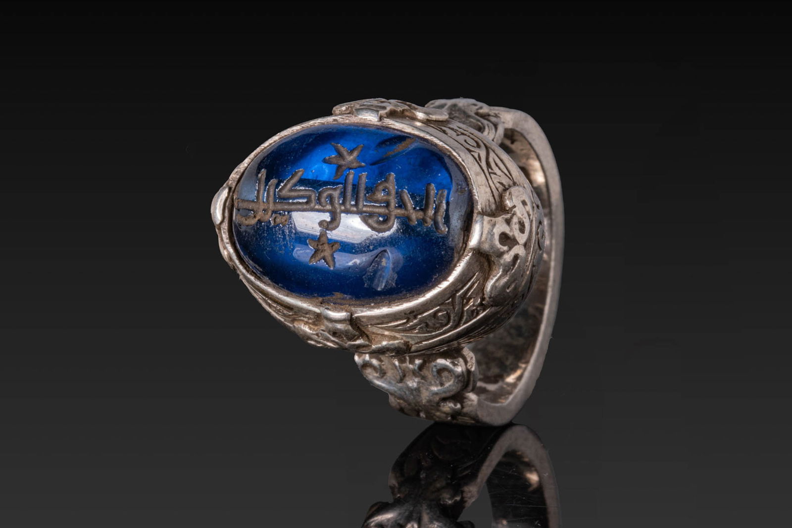 SASANIAN INSCRIBED BLUE INTAGLIO IN SILVER RING: Ca. AD 300 - 600. A cobalt blue oval intaglio engraved with a line of script, set in a silver ring with a high oval collet framed by a narrow rim and flanked at the quarters by small cusped bosses. Th