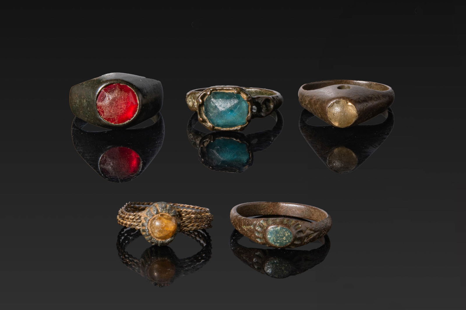 COLLECTION OF VIKING BRONZE RINGS WITH CABOCHONS: Ca. AD 700 - 1000. A collection of five bronze finger rings, including, from left to right: a heavy ring with a circular bezel set with a red cabochon; a ring with an octagonal bezel set with a blue g