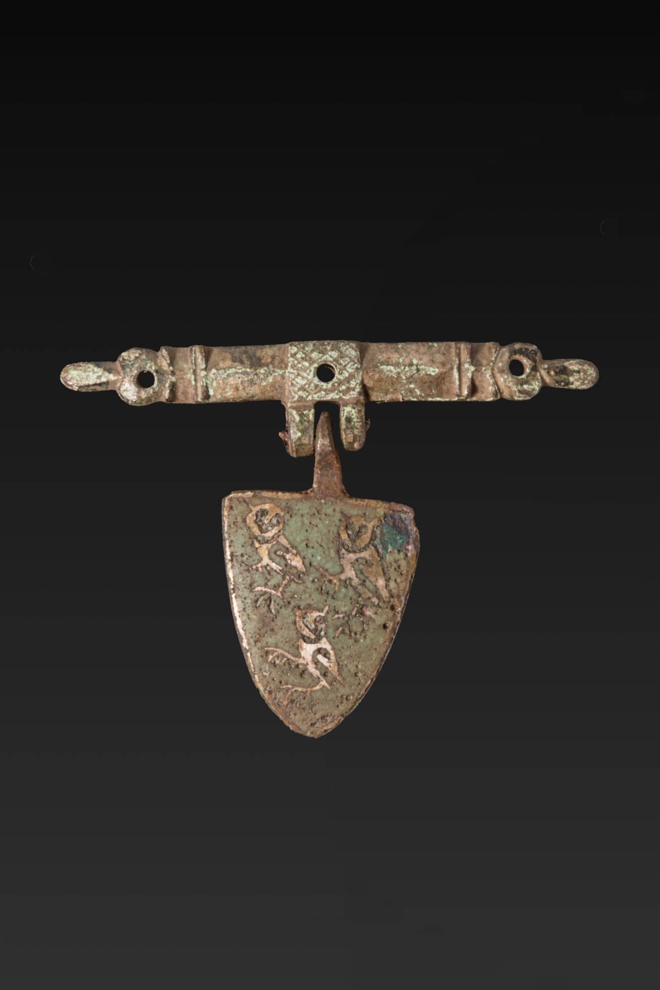BRITISH MEDIEVAL COPPER ALLOY HORSE HARNESS PENDANT WITH THREE OWLS: Ca. AD 1250 - 1400. A copper alloy horse harness pendant, with a transverse bar pierced at three points for attachment and a central hinge supporting a shield-shaped pendant. The shield is enamelled g