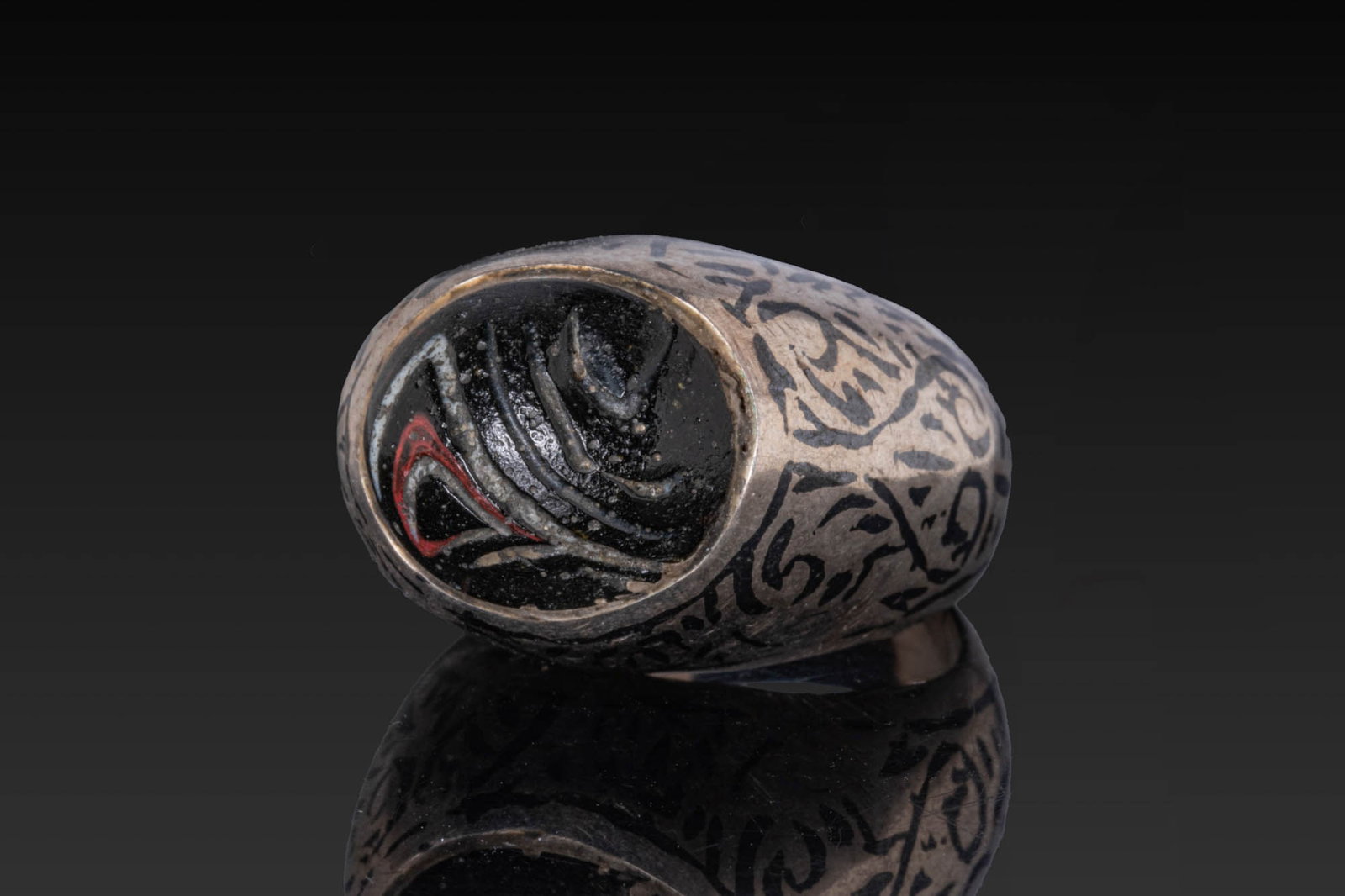 OTTOMAN OR QAJAR SILVER NIELLO RING: Ca. AD 1800 - 1900. A silver ring with a high bezel holding a dark glass insert with swirling lines inlaid with white and red. The upper hoop is decorated with niello in a dense scheme of scrolls and