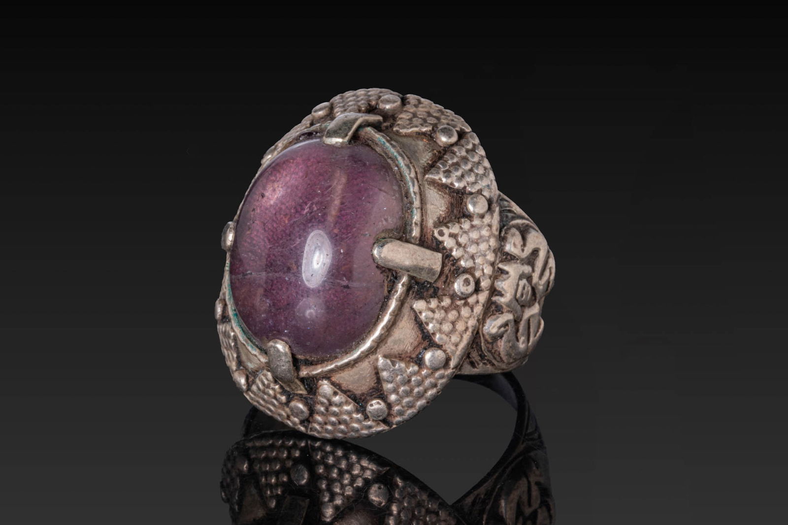OTTOMAN OR QAJAR SILVER RING WITH AMETHYST CABOCHON: Ca. AD 1800 - 1900. A silver ring with a flaring flat-section hoop supporting a high oval mount, set with an amethyst cabochon held by short prongs at the cardinal points. The mount is encircled by a
