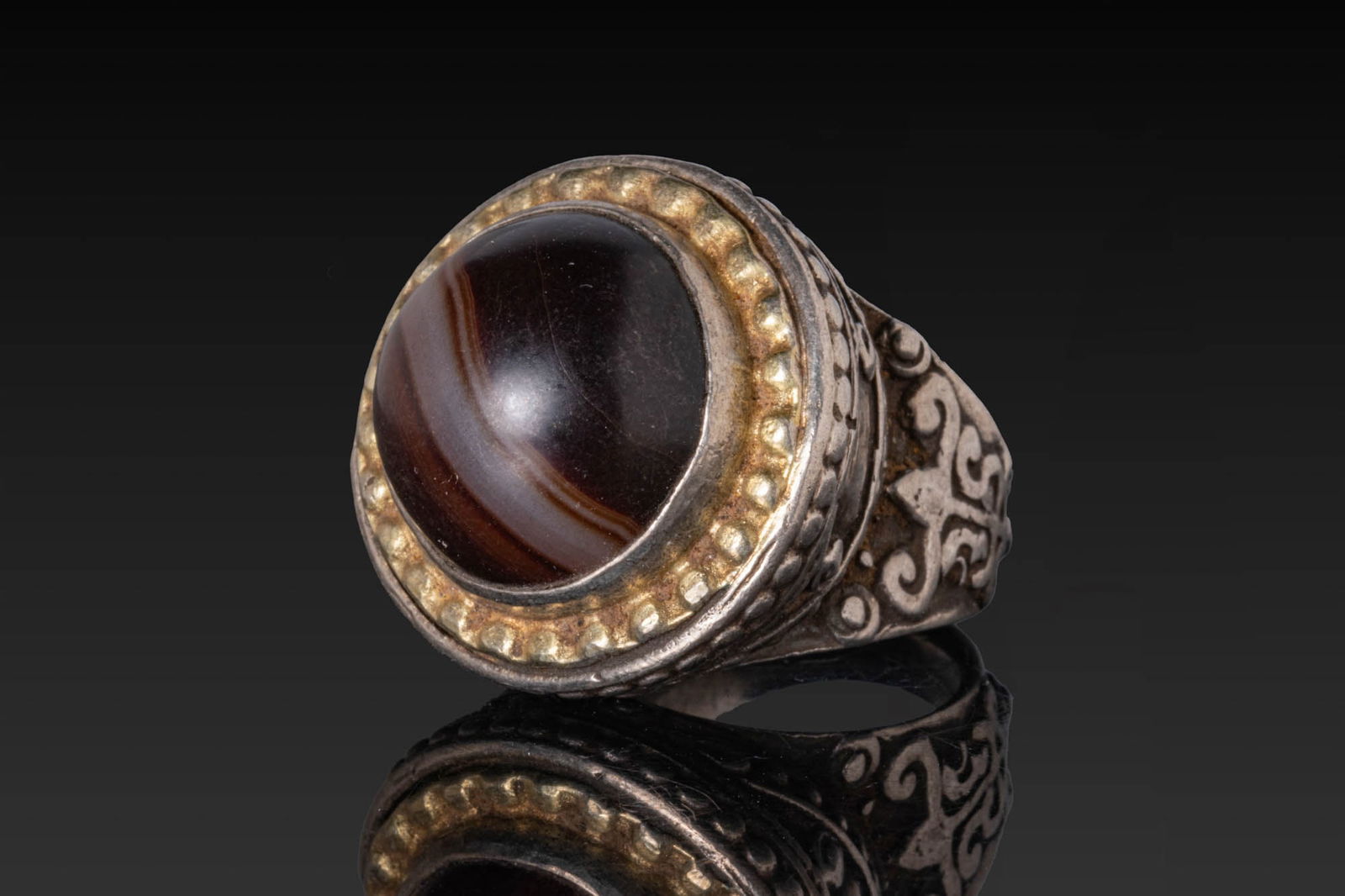 OTTOMAN OR QAJAR SILVER-GILT RING WITH BANDED AGATE CABOCHON: Ca. AD 1800 - 1900. A silver ring with a broad hoop supporting a high circular bezel set with a banded agate cabochon in a gilt frame, the shoulders decorated with scrollwork in low relief. Size: D:17