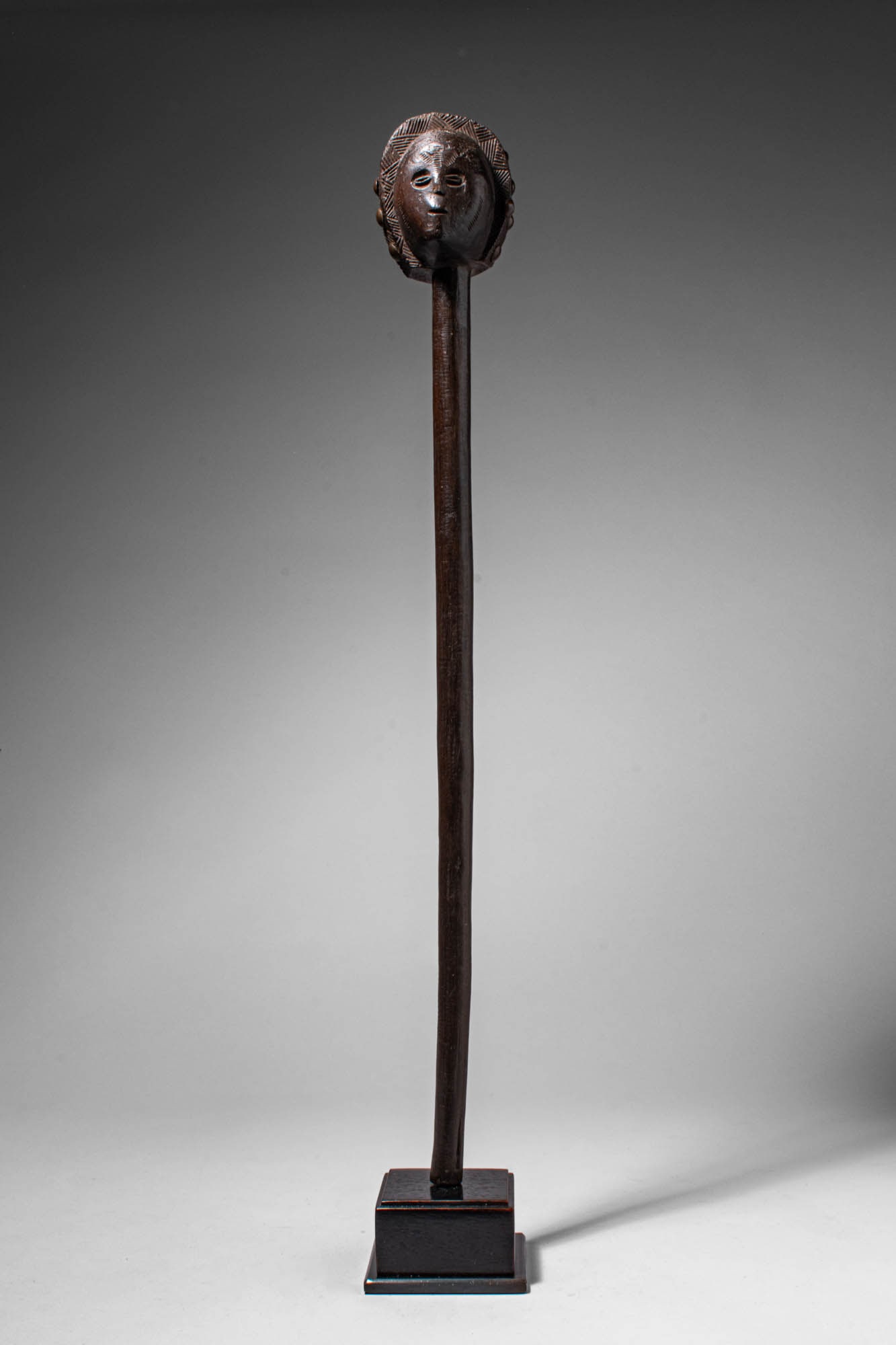 CHOKWE WOODEN STAFF: Angola, Ca. late 19th - early 20th century AD. >A slender staff surmounted by a carved globular head with stylised facial features, including small almond-shaped eyes, a straight nose, and a small mou