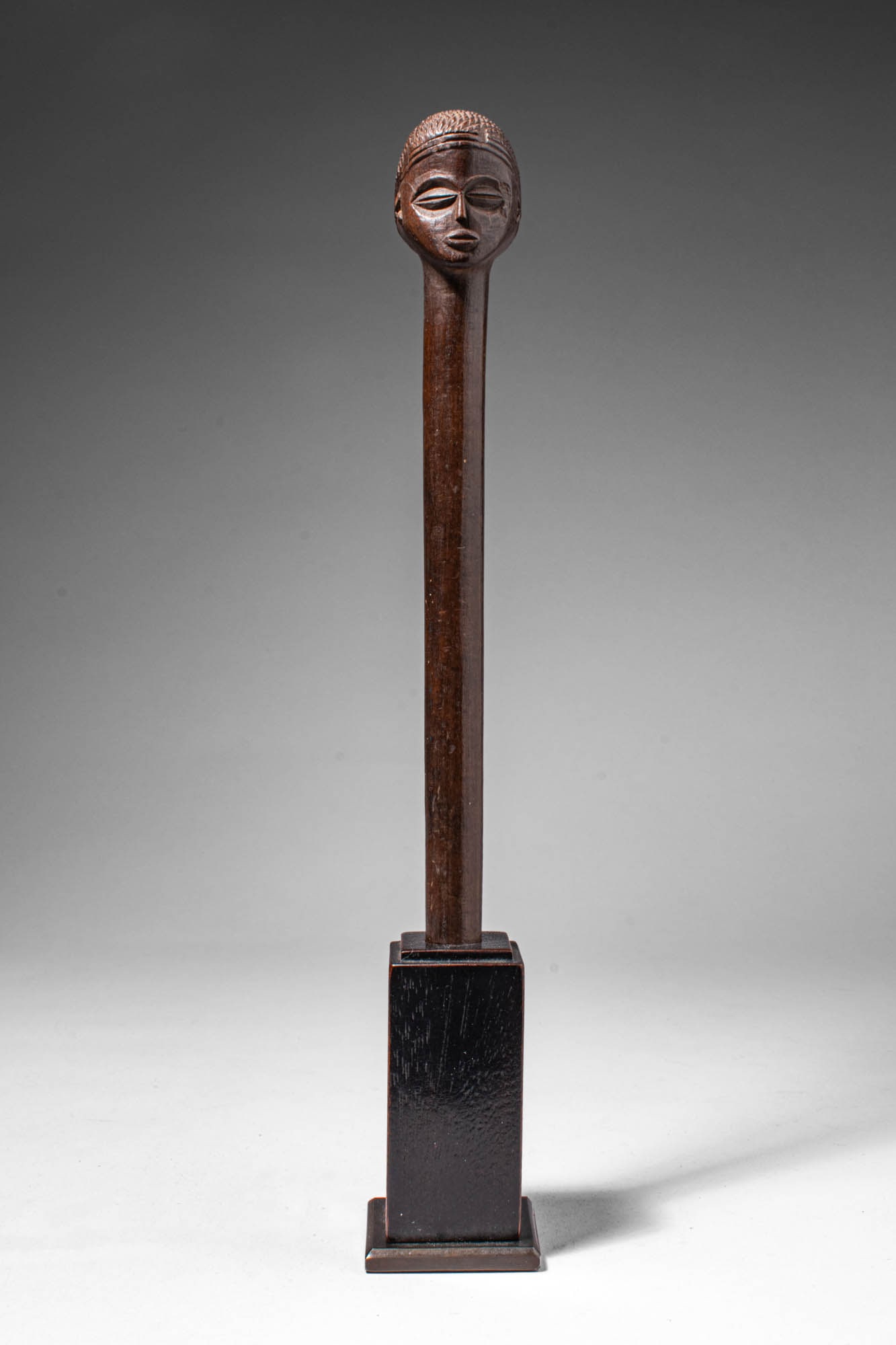CHOKWE WOODEN STAFF: Angola, Ca. late 19th - early 20th century AD. >A slender cylindrical staff surmounted by a carved globular head with stylised facial features, including almond-shaped eyes, a straight nose, and parte