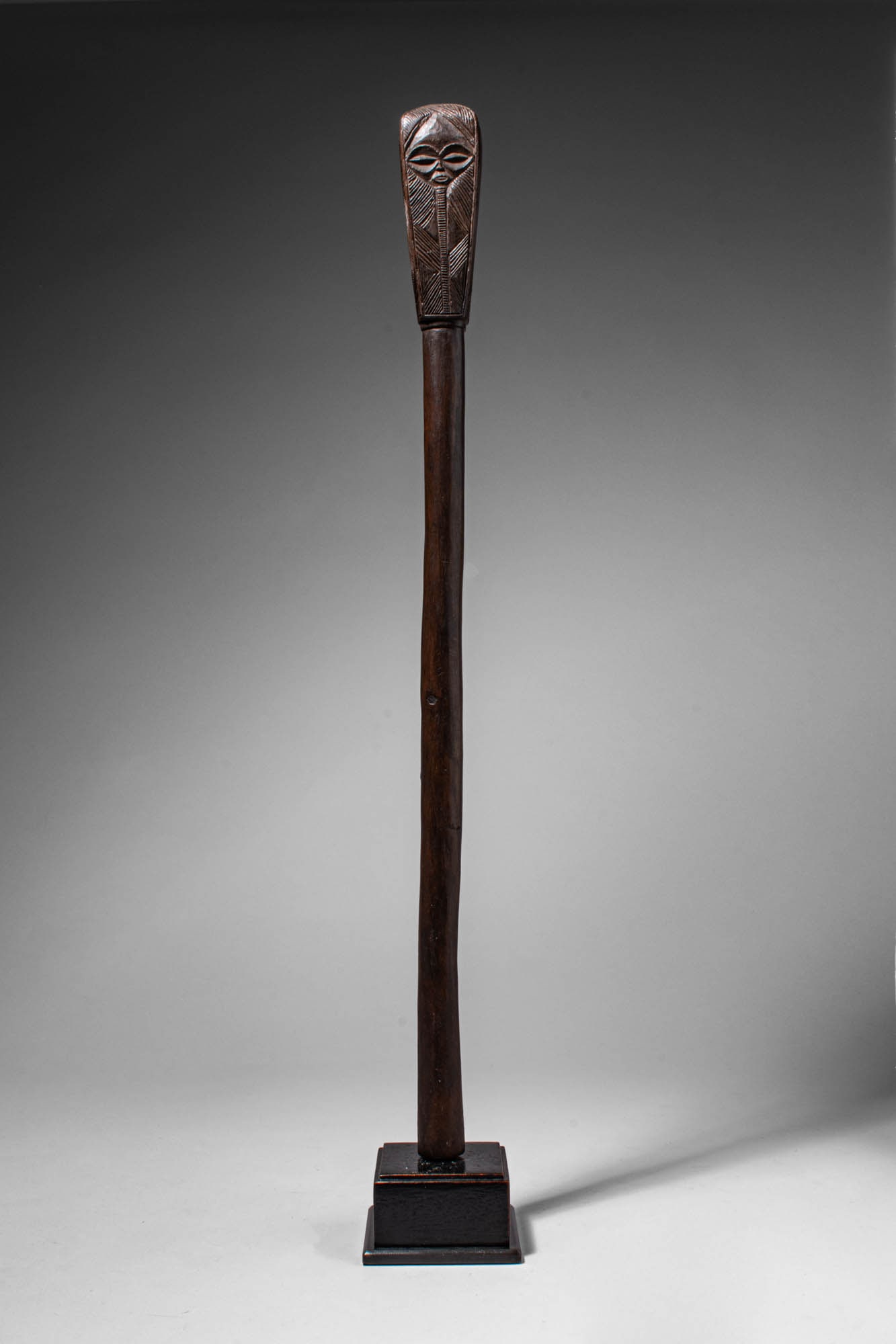 CHOKWE WOODEN STAFF: Angola, Ca. late 19th - early 20th century AD. >A long slender staff, surmounted by a carved rectangular head with stylised facial features, including almond-shaped eyes, a small mouth, and incised li