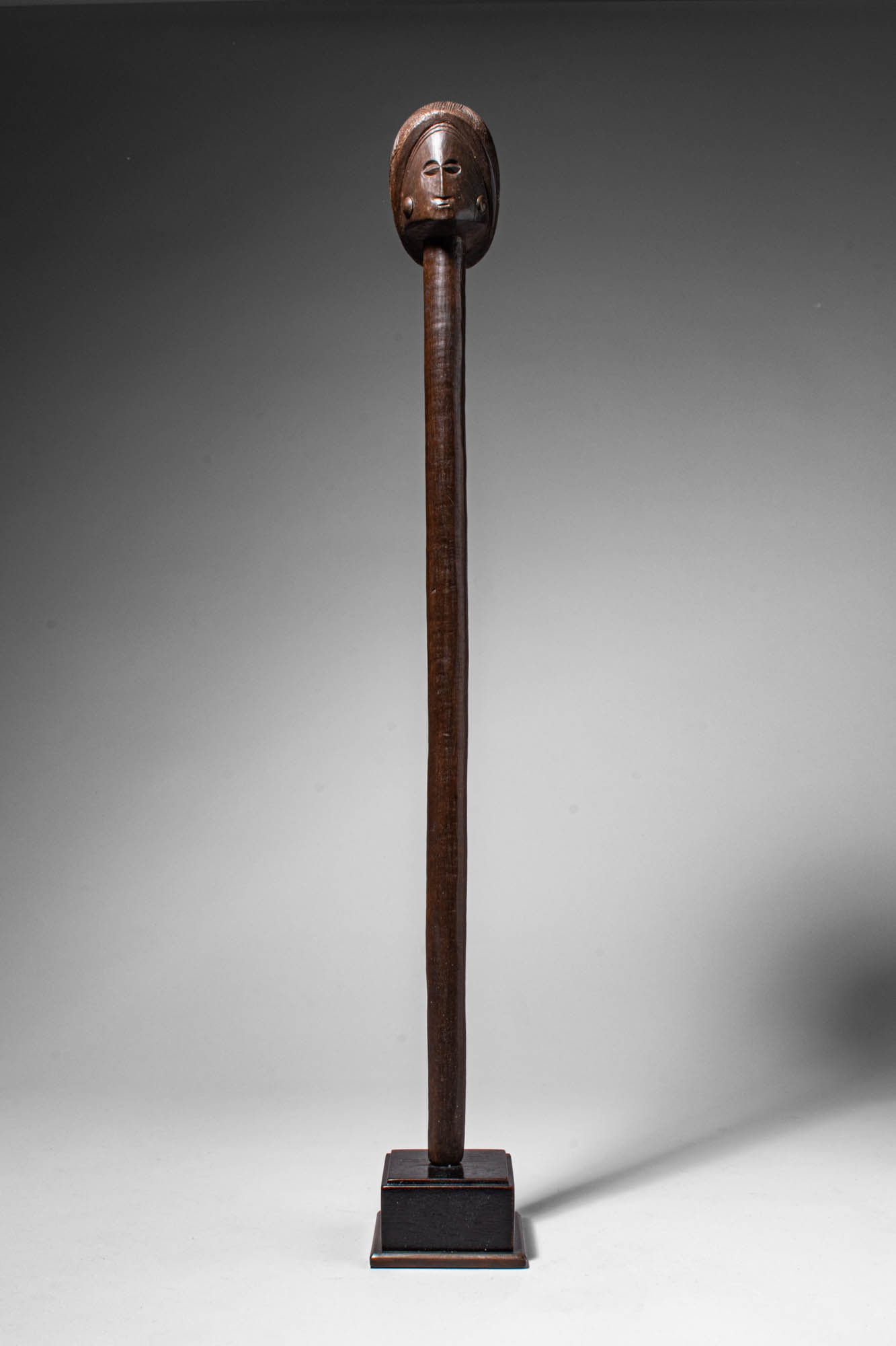 CHOKWE WOODEN STAFF: Angola, Ca. late 19th - early 20th century AD. >A slender staff surmounted by a carved globular head with stylised facial features, including almond-shaped eyes, a straight nose, and a small mouth. Th