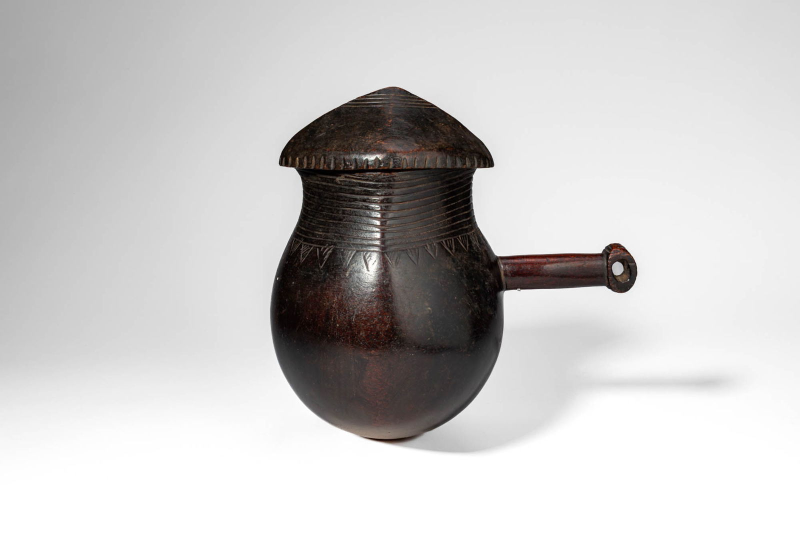 ZAIRE WOODEN SHI MILK POT AND LID: Ca. 20th century AD. >A wooden milk pot of globular form with a slightly tapering neck, the vessel carved from a single piece of wood and fitted with a conical domed cover. The body is decorated with
