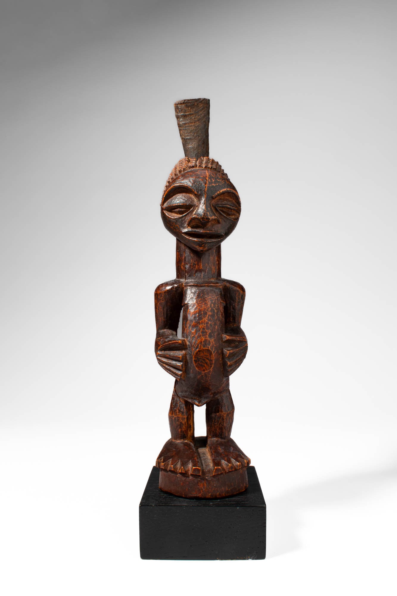 SONGYE WOODEN FIGURE: Ca. 20th century AD. >A standing Songye figure carved with compact proportions, the sculpture features a rounded head with large almond-shaped eyes, a small mouth, and a prominent vertical coiffure ri
