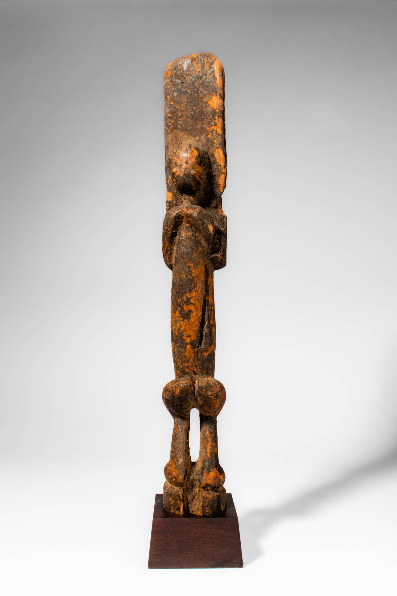DOGON NOMMO FIGURE: Ca. early 20th century AD. >A wooden Dogon Nommo figure carved as a highly elongated standing figure, featuring a long rectangular head and narrow torso. The arms are bent at the elbows and hands rest