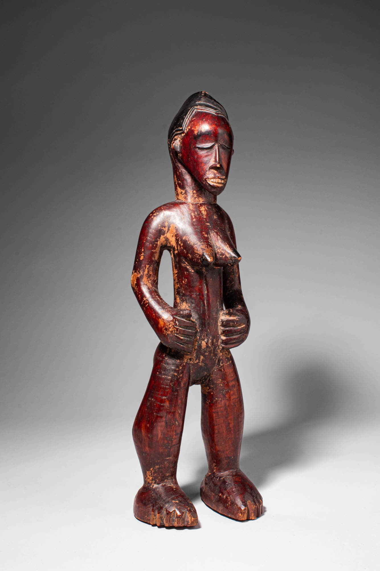 BEMBE WOODEN STANDING FIGURE: Ca. late 19th - early 20th century AD. > A Bembe wooden standing figure carved with slightly flexed legs and hands placed across the abdomen, the sculpture displays the elongated proportions character