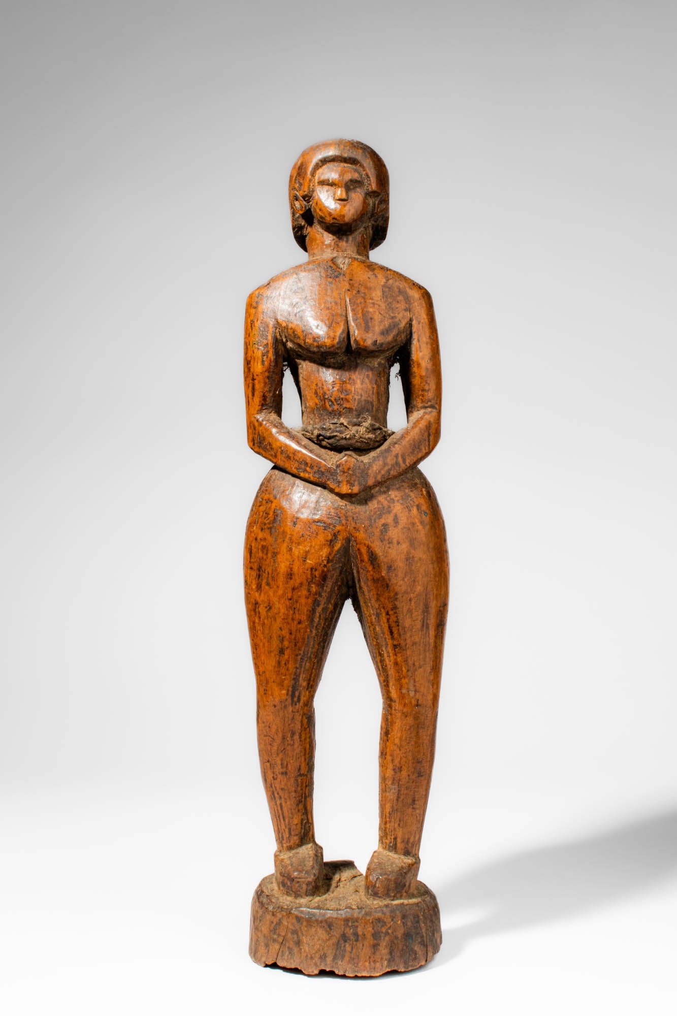 TANZANIAN WOODEN FEMALE FIGURE: Ca. mid 20th century AD or earlier. >A Tanzanian wooden female figure with naturalistic proportions, the sculpture depicts a woman standing upright with her arms bent and hands joined at the abdomen.