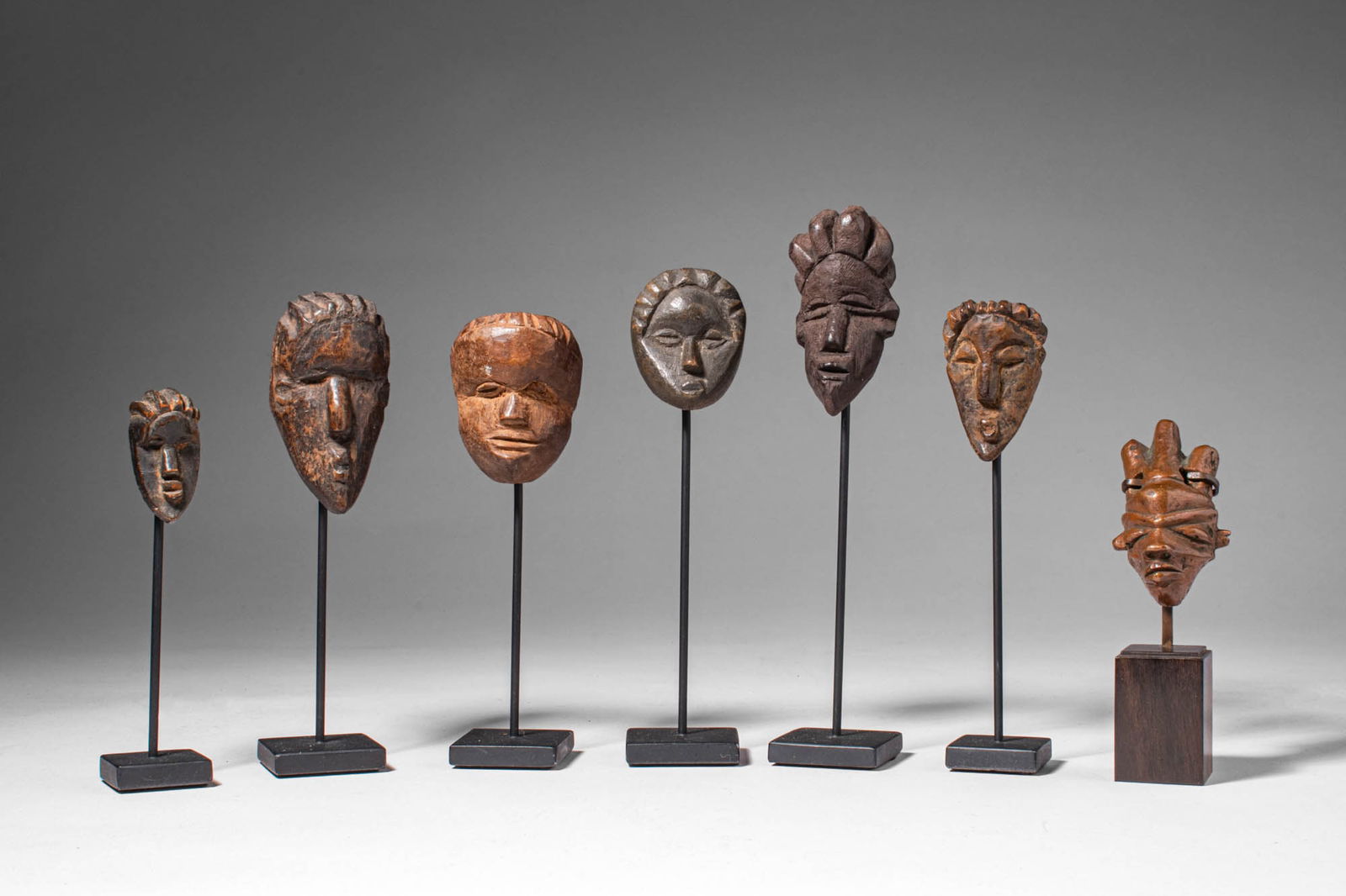 GROUP OF MINIATURE MASKS: Ca. 19th - 20th century AD. >A group of six carved wooden miniature masks and one cast metal example (bronze or brass), each depicting stylised facial features characteristic of various West and Centr