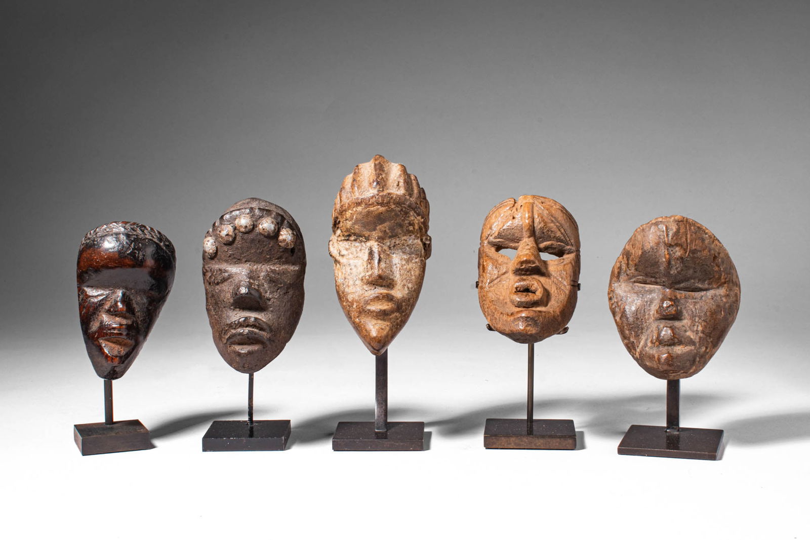GROUP OF DAN PASSPORT MASKS: Ca. 19th - 20th century AD. >A group of five miniature carved wooden masks with elongated oval faces and stylized facial features, including almond-shaped or slit eyes, pronounced noses and incised co