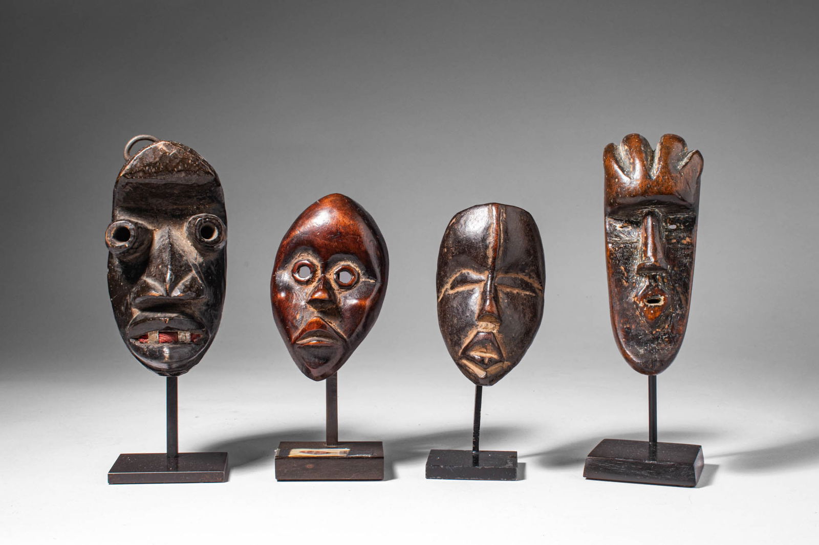 GROUP OF DAN PASSPORT MASKS: Ca. 19th - 20th century AD. >A group of four miniature carved wooden masks with elongated oval forms and stylized facial features, including circular or almond-shaped eyes, pronounced noses and incise