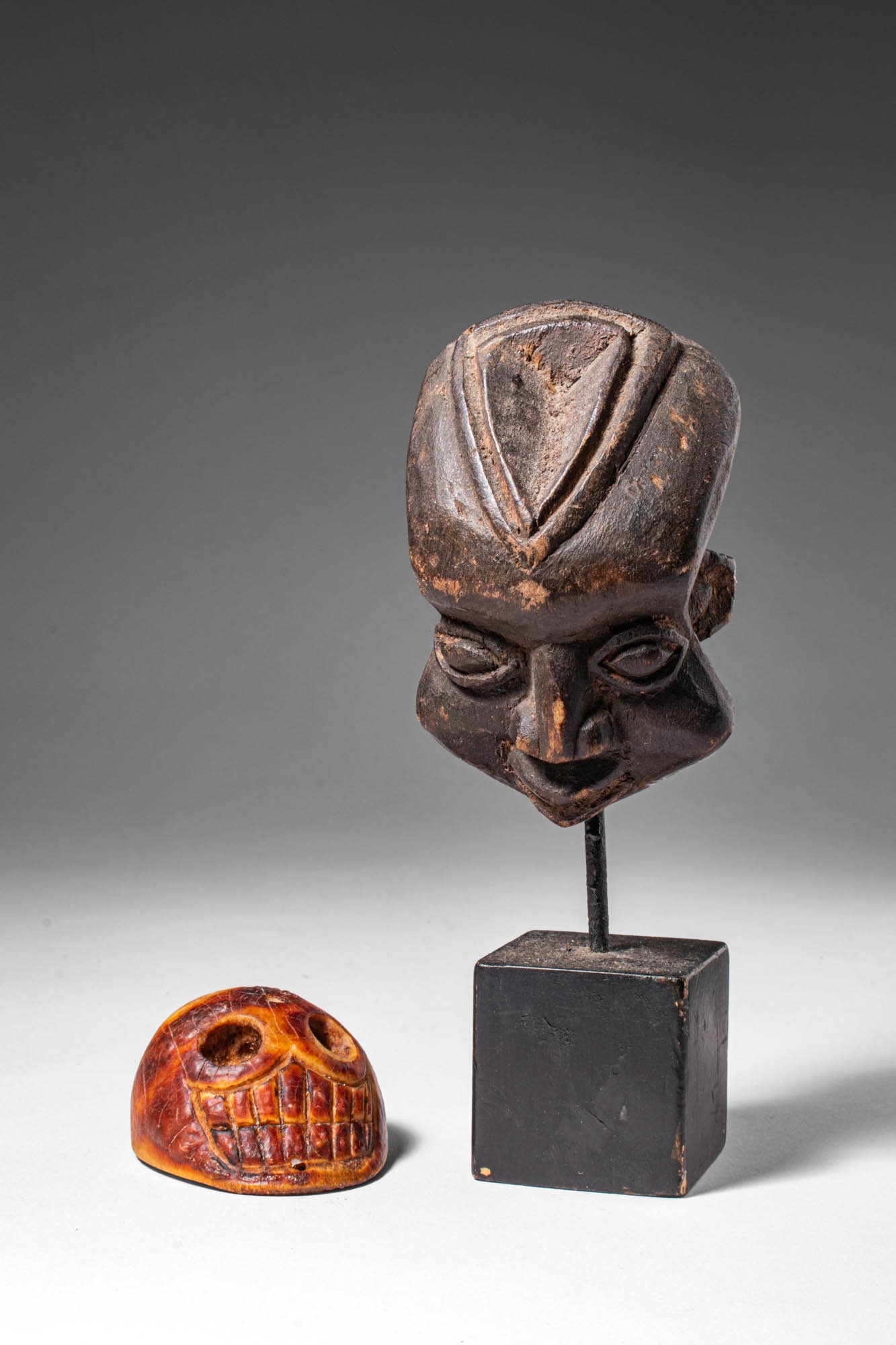 KAPALA SKULL BEAD AND AFRICAN WOODEN MASK: Ca. 20th century AD. >A small group comprising a kapala bead and a carved African wooden mask head mounted for display.The kapala bead, carved from bone, takes the form of a stylised skull, with deepl