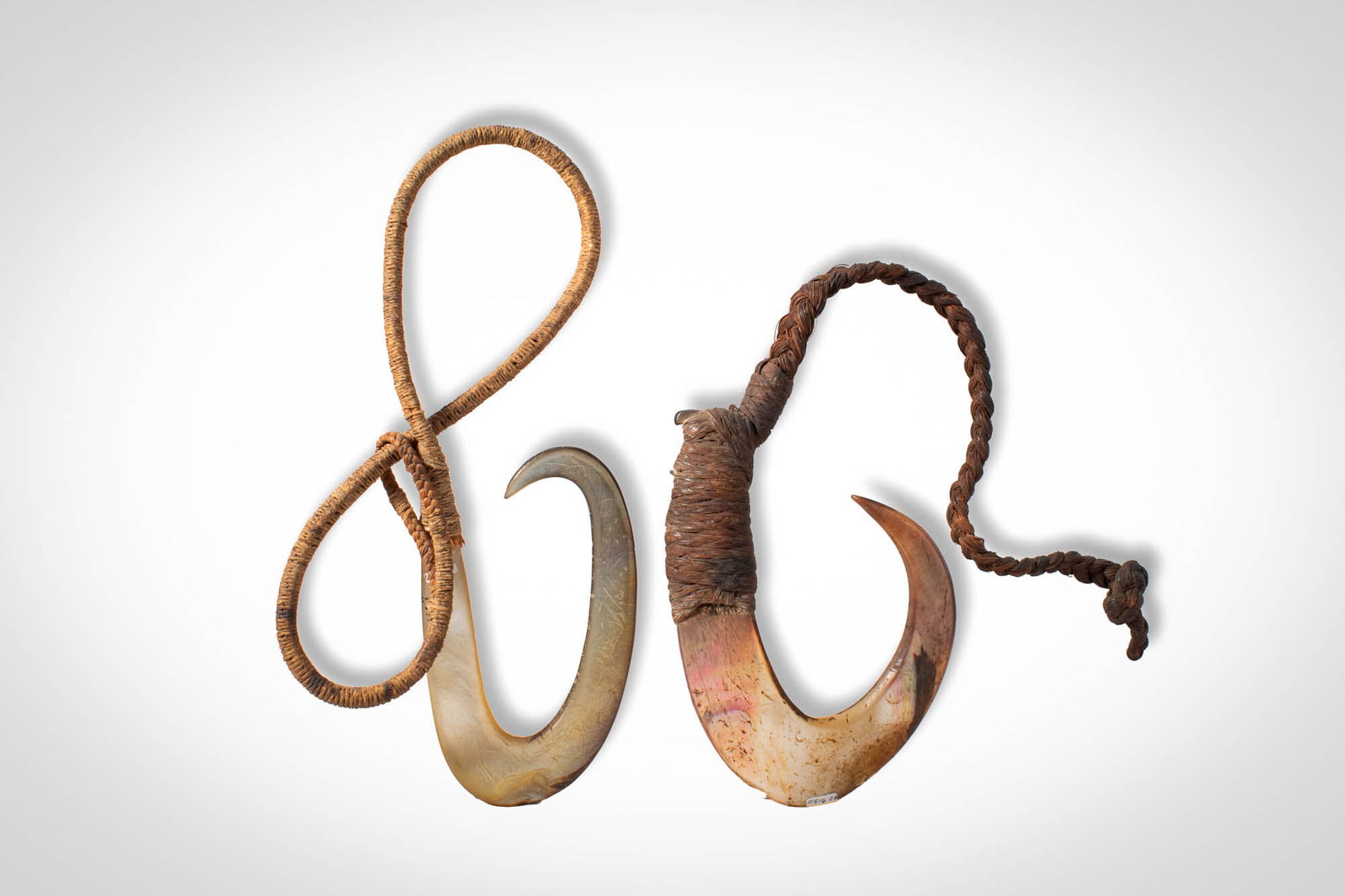 TWO TAHITI ISLAND FISH HOOKS: Ca. 19th - 20th century AD. >A group of two fish hooks carved from shell, each formed with a broad, curved body tapering to sharply pointed terminals. Both examples retain a long braided fibre cord at
