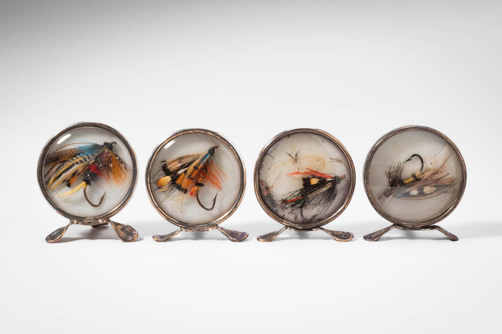 MATCHED SET OF FOUR EDWARDIAN SILVER NOVELTY MENU CARD HOLDERS (GREY & CO): Ca. early 20th century AD. >A group of four silver novelty menu card holders. Each circular holder is formed with a domed front enclosing a carefully arranged fishing fly. The four examples display di