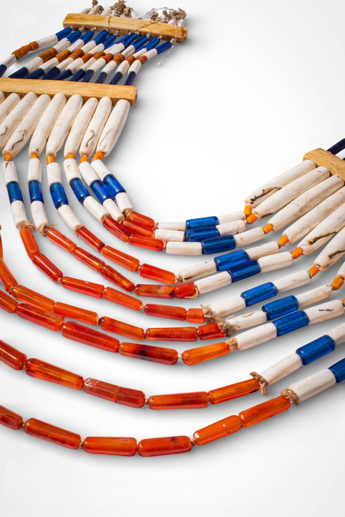 INDIAN NAGALAND POLYCHROME NECKLACE: Ca. 19th - 20th century AD. >A necklace composed of multiple suspended strands of elongated carnelian, glass, and shell beads arranged in alternating bands of red, blue, and white. The strands are sec