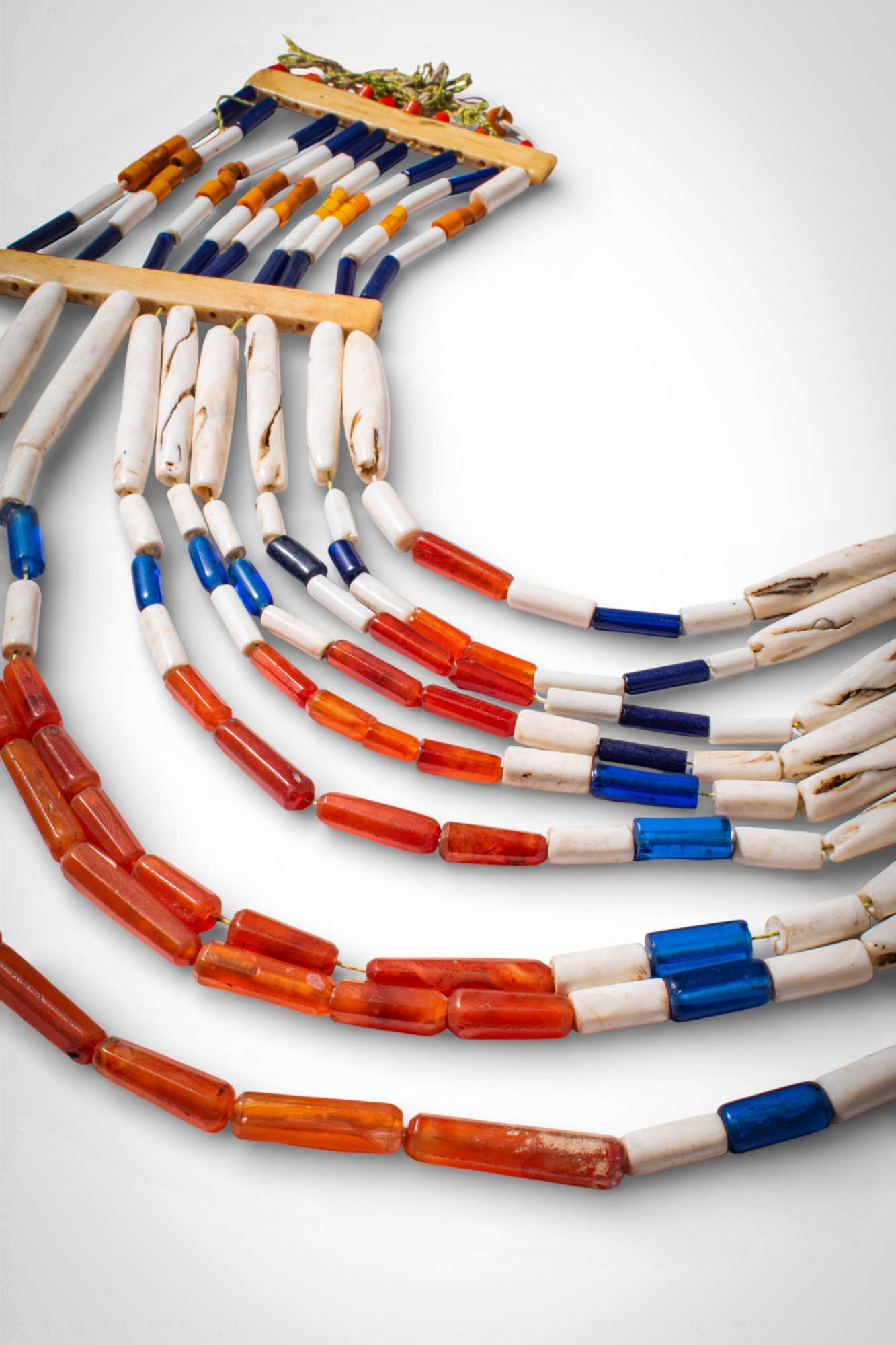 INDIAN NAGALAND POLYCHROME NECKLACE: Ca. 19th - 20th century AD. >A necklace composed of multiple suspended strands of elongated carnelian, glass, and shell beads arranged in alternating bands of red, blue, and white. The strands are sec