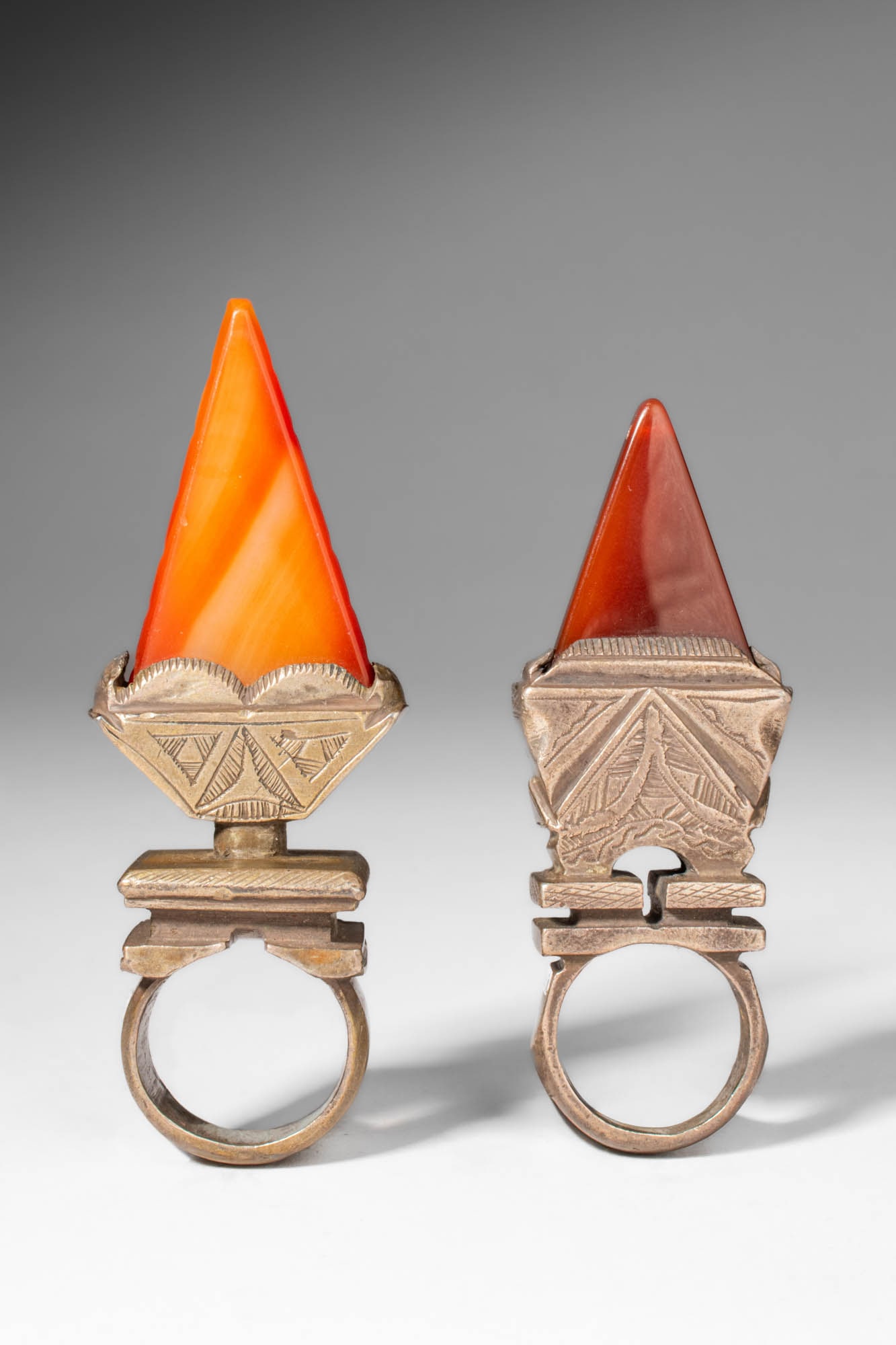 TWO TUAREG RINGS: Ca. 20th century AD. >A group of two rings, each set with a tall, conical carnelian element rising above a geometric mount. The mounts are engraved with linear and triangular motifs characteristic of