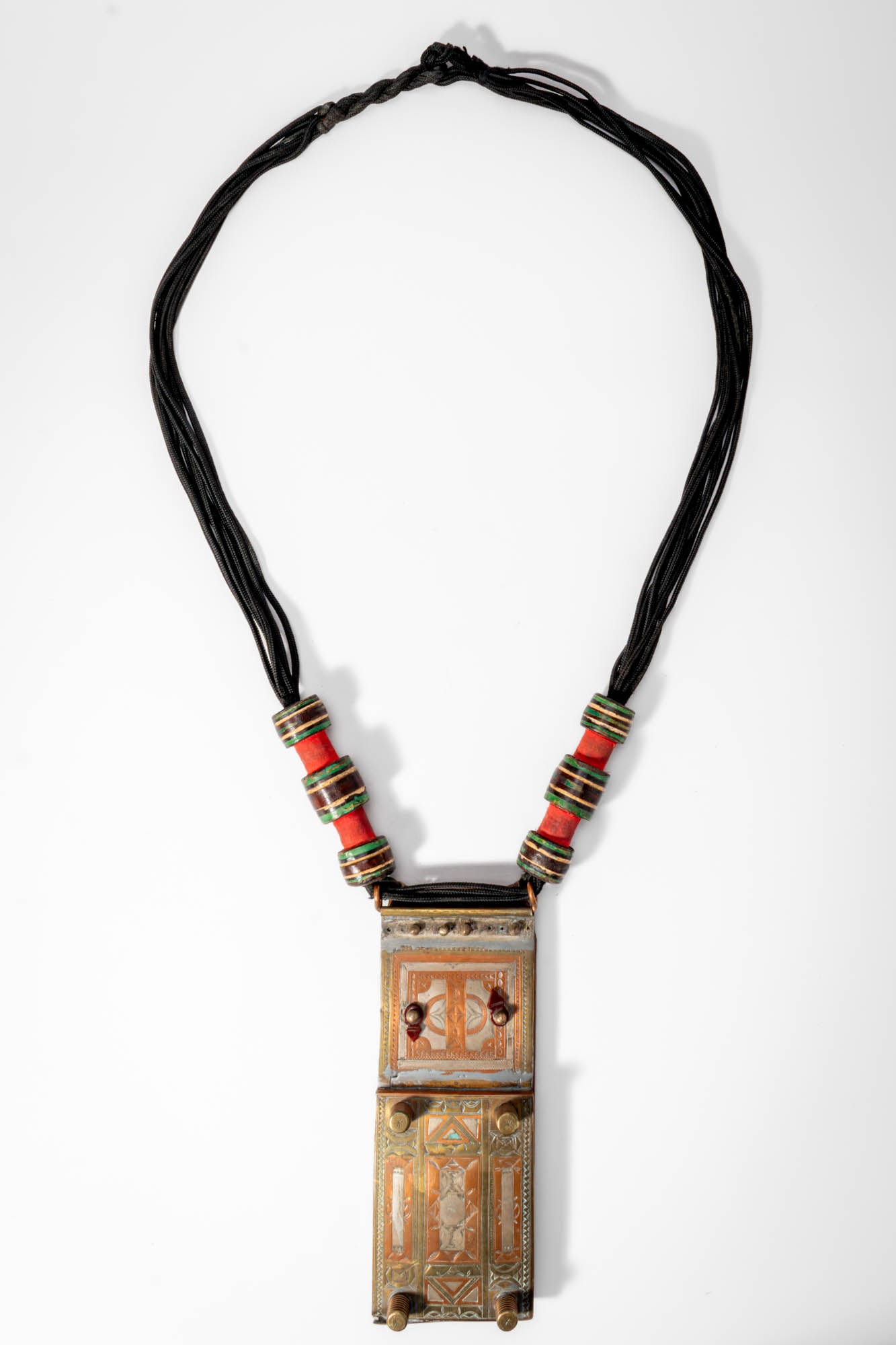 TUAREG METAL POUCH: Ca. 20th century AD. >A Tuareg metal pouch pendant suspended from a multi-strand leather cord necklace. The rectangular container is decorated with finely engraved geometric motifs and panels of contr