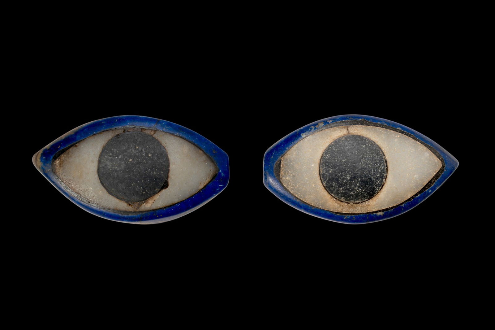 EGYPTIAN GLASS MUMMY MASK EYES: Ca. 2200 - 1800 BC. A pair of inlaid eyes, each formed from a flat-backed, almond-shaped glass plaque with a white stone insert for the sclera and a black stone disc for the iris. Size: 25-25mm x 44-4