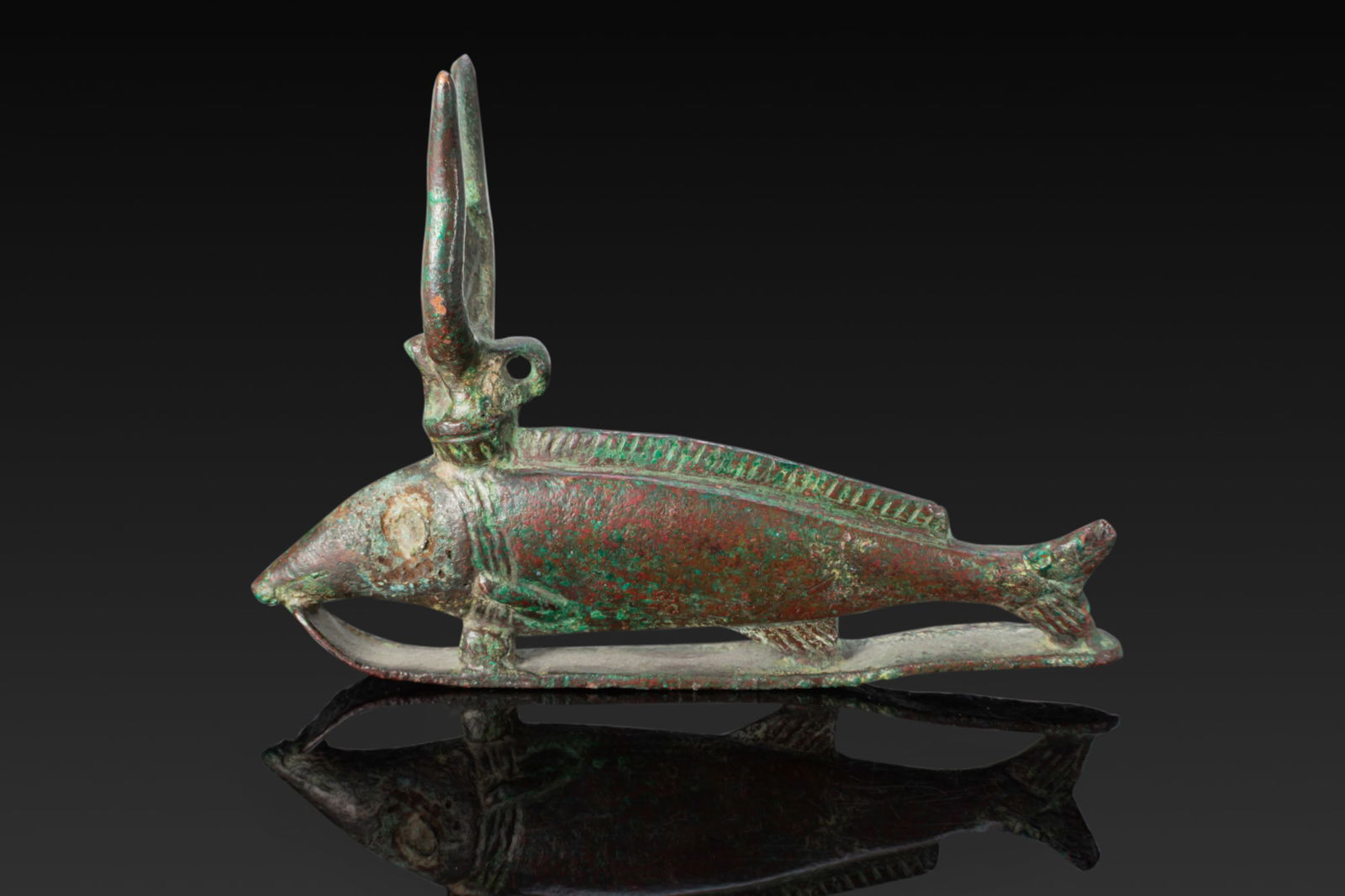 EGYPTIAN BRONZE OXYRHYNCHUS FISH AMULET: Late Period, Ca. 664 - 332 BC. A bronze amulet cast in the form of an oxyrhynchus fish, set on a long flat base strip, the head tapering to a pointed snout and the body with a raised dorsal fin and a