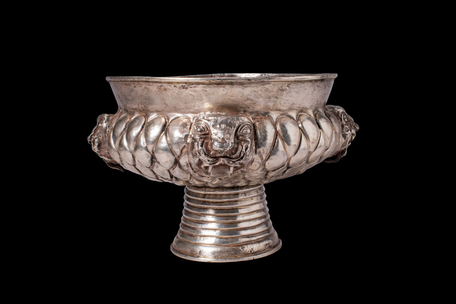 STUNNING GREEK PERIOD SILVER FOOTED BOWL WITH LION PROTOMES: Ca. 600 - 400 BC. A silver footed bowl with a deep, rounded body on a ribbed pedestal foot. The exterior is worked in repouss? with an overlapping scale pattern, interrupted at the cardinal points by