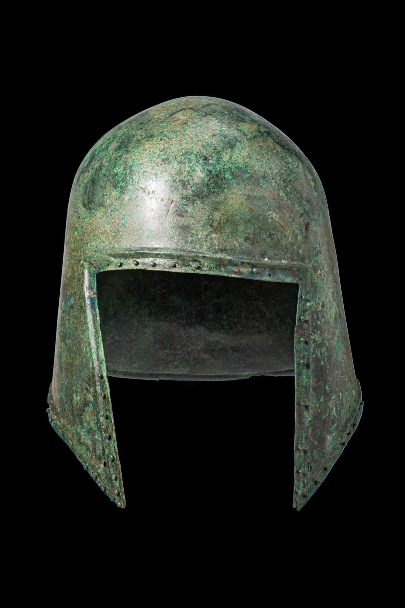 LARGE ANCIENT GREEK ILLYRIAN BRONZE HELMET: Ca. 600 - 500 BC. A bronze helmet with a rounded crown and a broad rectangular face opening, the margins reinforced and pierced with a line of rivet holes. The sides descend into long cheekpieces narr