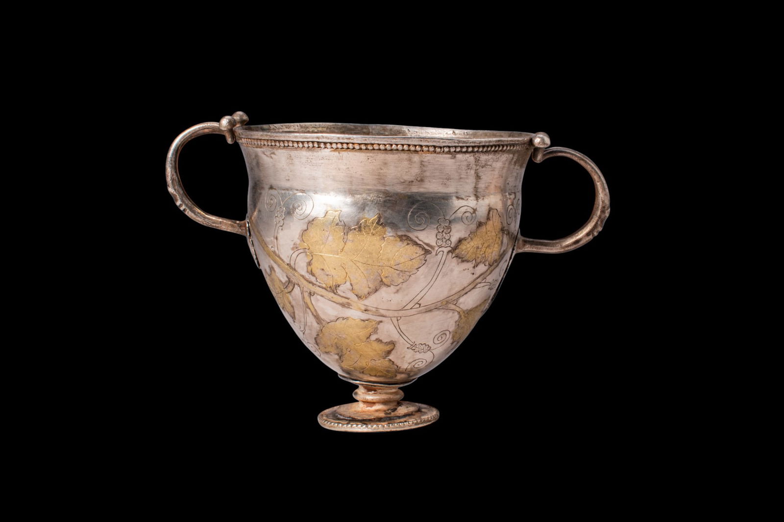 STUNNING LATE ROMAN SILVER-GILT SKYPHOS WITH VINE LEAVES MOTIF: Ca. AD 300 - 500. A silver-gilt skyphos on a low spreading foot, the body rising from a short stem to a flared rim with a beaded band below the lip. Two opposed bifurcated handles are applied to the u