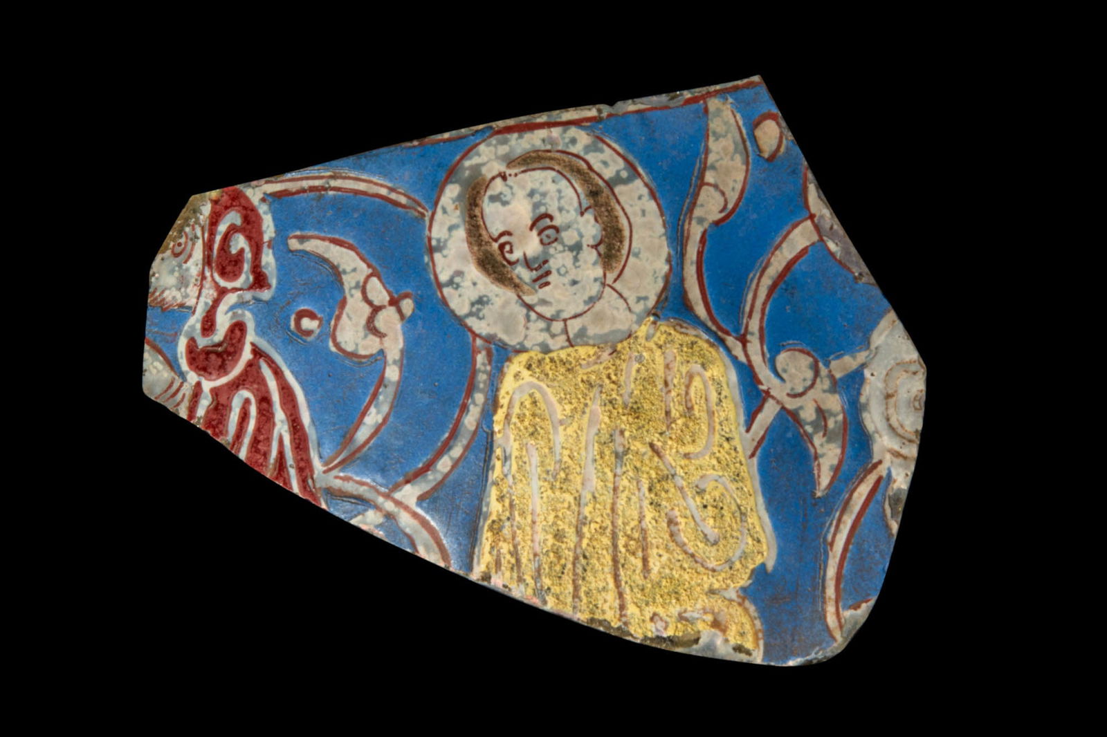 VERY RARE SYRO-FRANKISCH GLASS FRAGMENT: Ca. AD 1260. A polychrome glass fragment with a blue ground, featuring figural composition outlined in red. The central figure, depicted frontally, has a rounded face with large eyes, a straight nose,