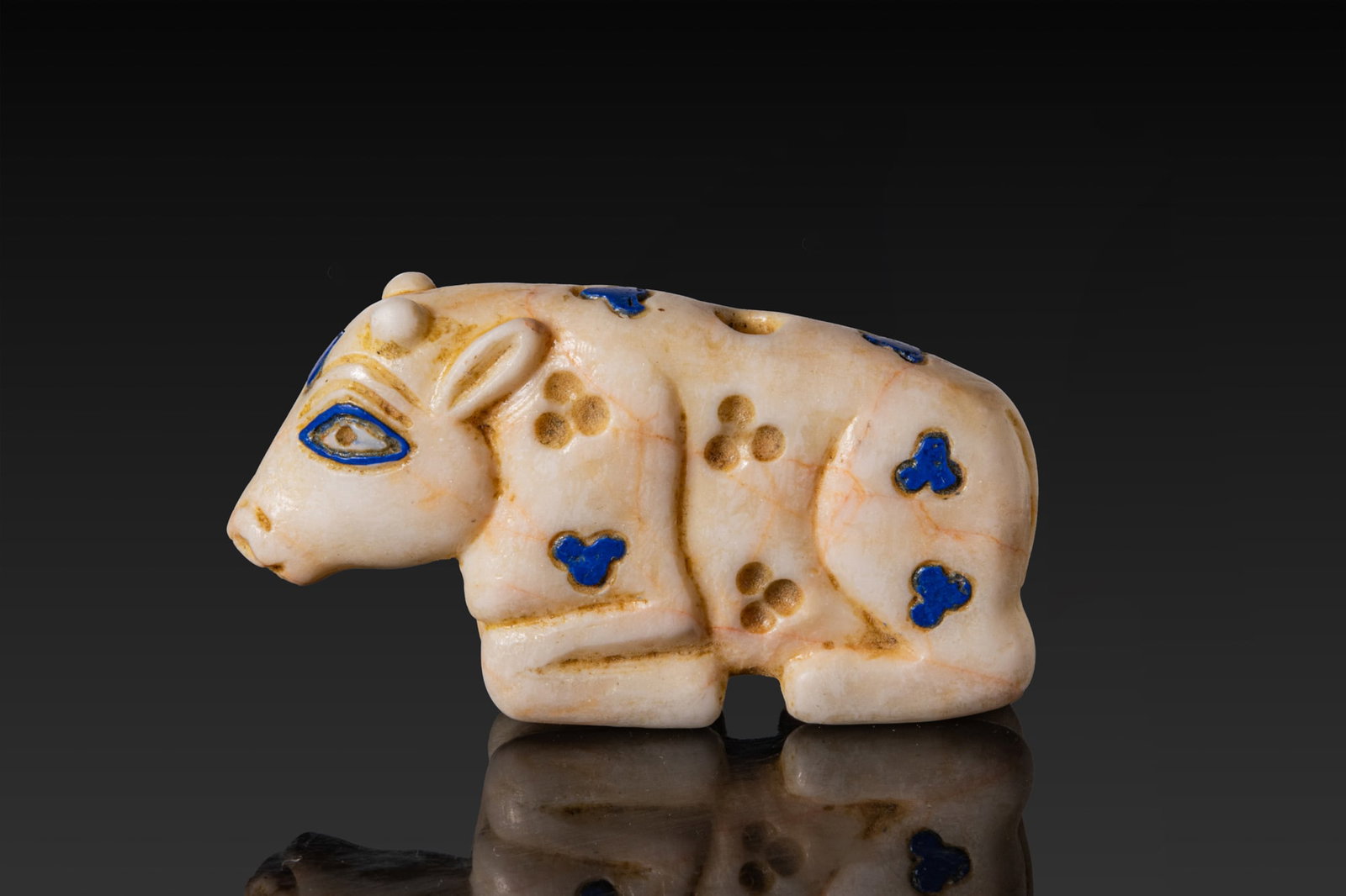 MESOPOTAMIAN MARBLE AND LAPIS LAZULI RECUMBENT CALF: Late Uruk - Jemdet Nasr, Ca. 3300 - 2900 BC. A marble figure of a recumbent calf, carved with folded legs beneath a rounded body and a short tail drawn down the haunch. The head is modelled with a rou