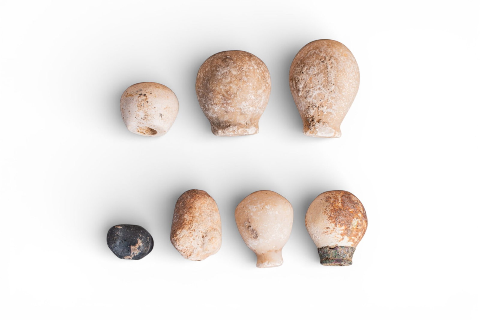 COLLECTION OF SUMERIAN STONE MACE HEADS: Ca. 2500 - 1800 BC. A collection of seven stone mace heads, each with a centrally drilled shaft, including, from left to right: two pear-shaped examples, one with a bronze collar and one tapering to a