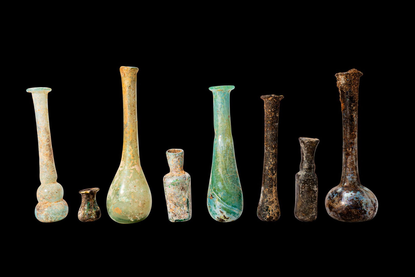 COLLECTION OF ROMAN GLASS UNGUENTARIA: Ca. AD 1 - 300. A collection of eight glass unguentaria including, from left to right: a tall vessel with a long cylindrical neck above a double-bulbed lower body; a miniature squat bottle with a roun