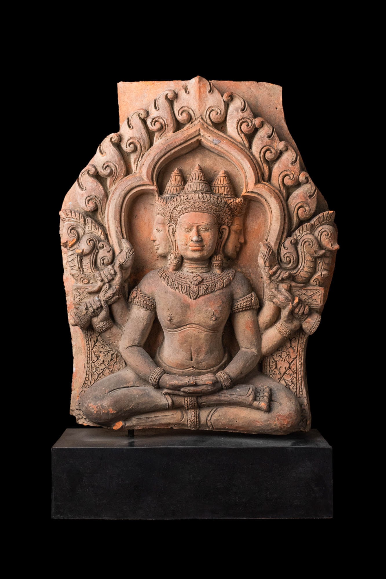 KHMER TERRACOTTA RELIEF OF A FOUR-ARMED AND THREE-HEADED DEITY (LIKELY BRAHMA): Possibly Angkor period, Ca. AD 1100 - 1300. A fine terracotta relief depicting a three-headed and four-armed deity, likely Brahma, seated within an elaborate architectural frame. The central face is r