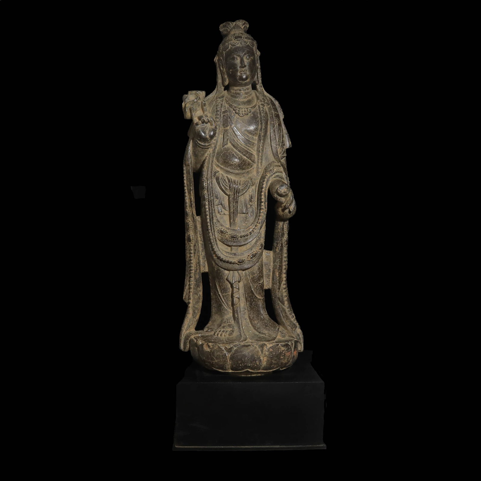 LARGE CHINESE TANG DYNASTY STONE FIGURE: Ca. AD 618 - 907. A large figure standing erect wearing a loosely draped pleated garment with beaded swags over the forearms, segmented hairstyle held with a diadem, right arm raised in gesture of rea