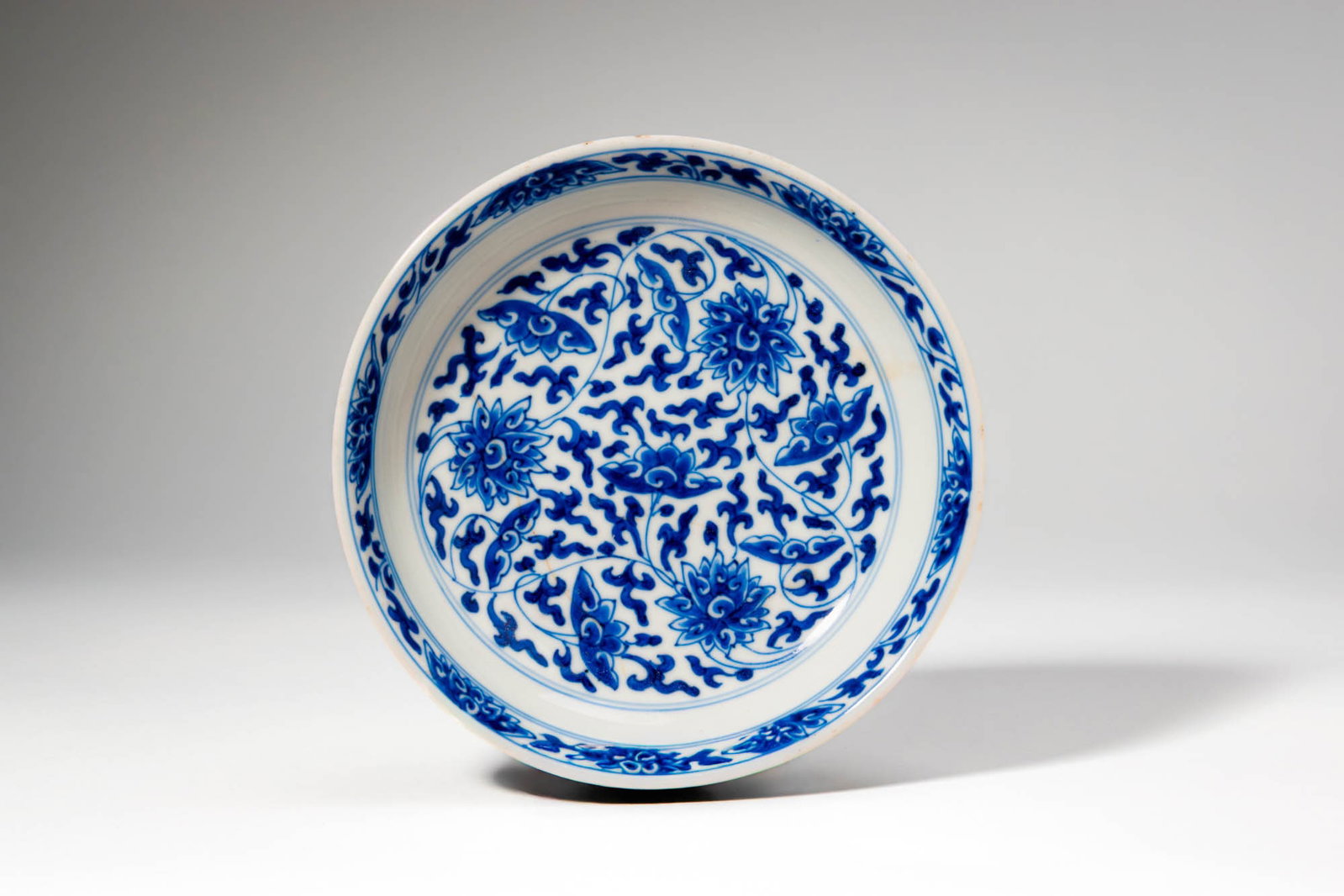 CHINESE BLUE AND WHITE LOTUS DISH, KANGXI MARK: Kangxi Period, Qing Dynasty, Ca. AD 1661 - 1722. >A finely potted blue and white porcelain dish of shallow rounded form, the interior decorated in rich cobalt-blue tones with a dense scrolling lotus d