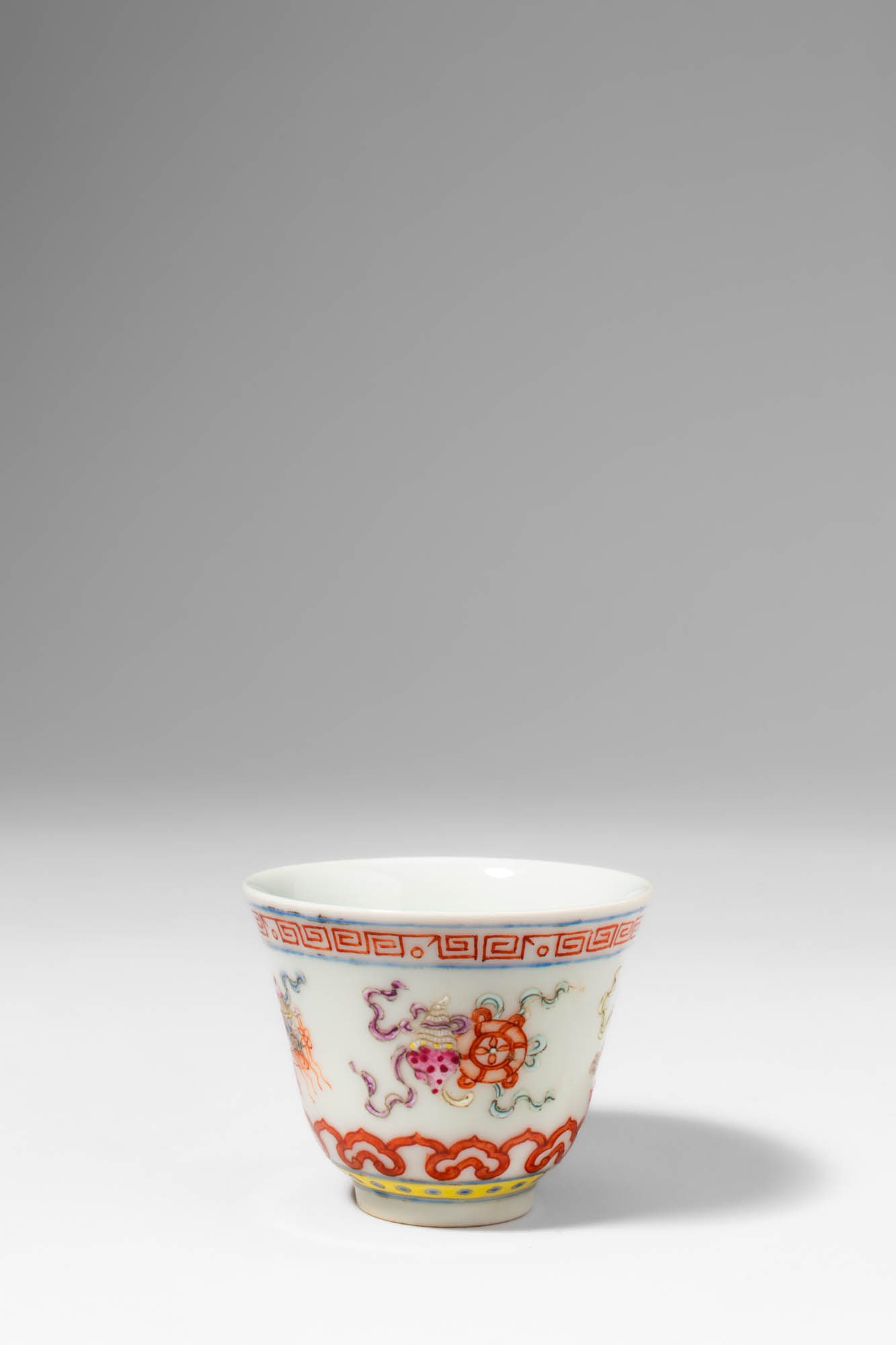 CHINESE IRON RED DECORATED CUP, DEPICTING THE EIGHT AUSPICIOUS SYMBOLS: . >A porcelain cup of slightly flared form, resting on a neatly trimmed foot, and decorated in a lively overglaze enamel palette dominated by iron-red.The exterior is adorned with elements of the Eigh