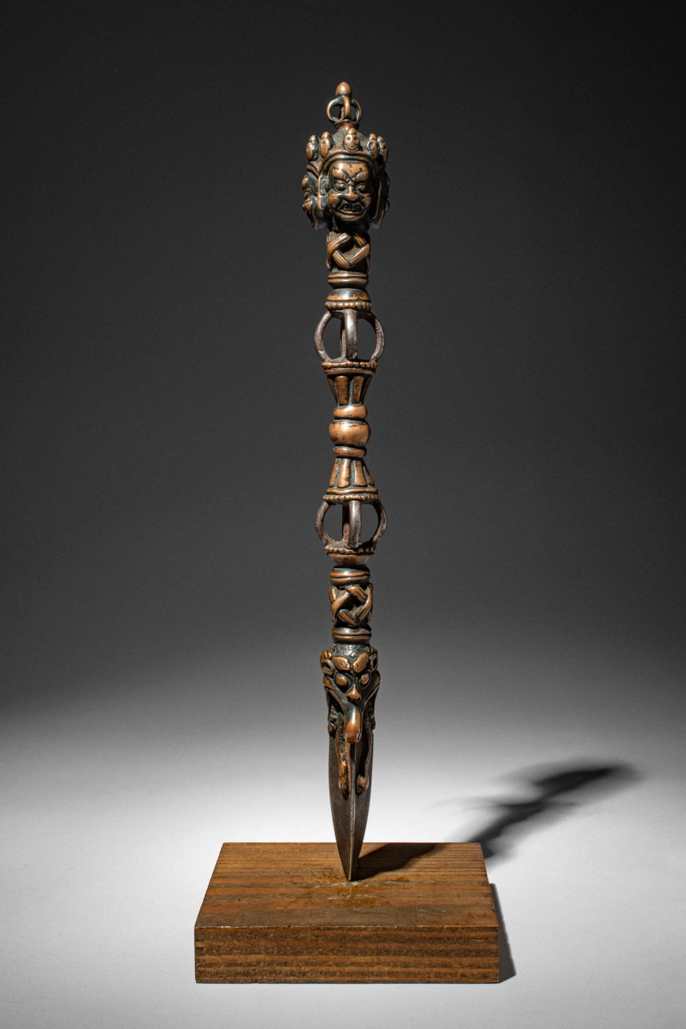 BRONZE AND CAST IRON PHURBU, TIBETAN OR NEPALESE: Ca. 16th century AD. >A finely cast ritual phurba (kila), composed of a handle and a trihedral blade, the shaft surmounted by a wrathful deity head beneath a foliate crown. The deity?s fierce expressi