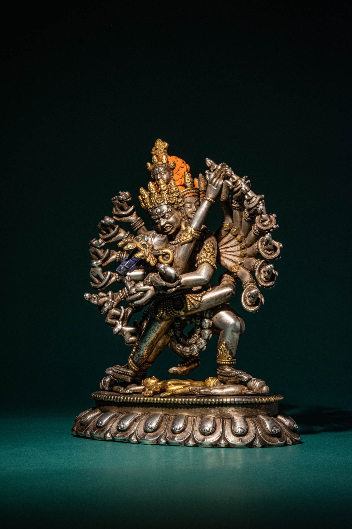 TIBETAN SILVER WITH PARCEL GILT FIGURE OF CHAKRASAMVARA: Ca. 18th-19th century AD. > A powerful silver and parcel-gilt figure of Chakrasamvara in union with his consort, standing on a lotus base over figures. Chakrasamvara, depicted with twelve arms, four h