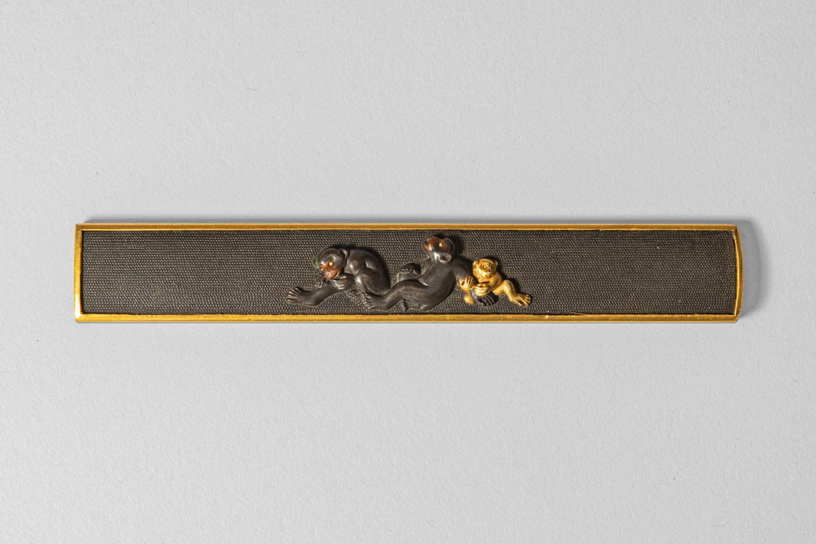 JAPANESE KOZUKA, SIGNED IWAMOTO: Edo Period, Ca. 19th century AD. A refined kozuka (small knife handle) featuring a lively depiction of two monkeys and a young one, finely rendered in shakudo and gold in high relief. The dark nanako 
