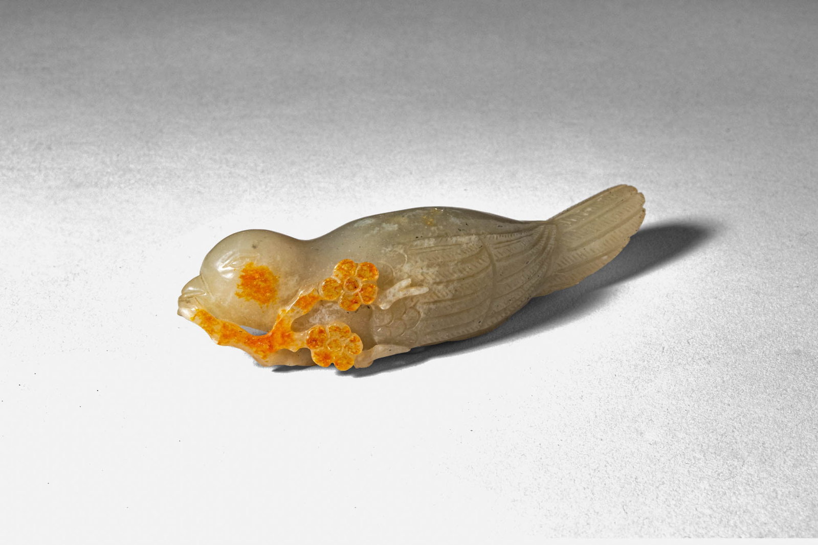 CHINESE PALE GREY JADE MAGPIE: Qing Dynasty, Ca. AD 1644 - 1912. >A delicately carved jade figure of a recumbent magpie, the bird shown with its body elongated and head slightly lowered as it grasps a flowering spray in its beak. T
