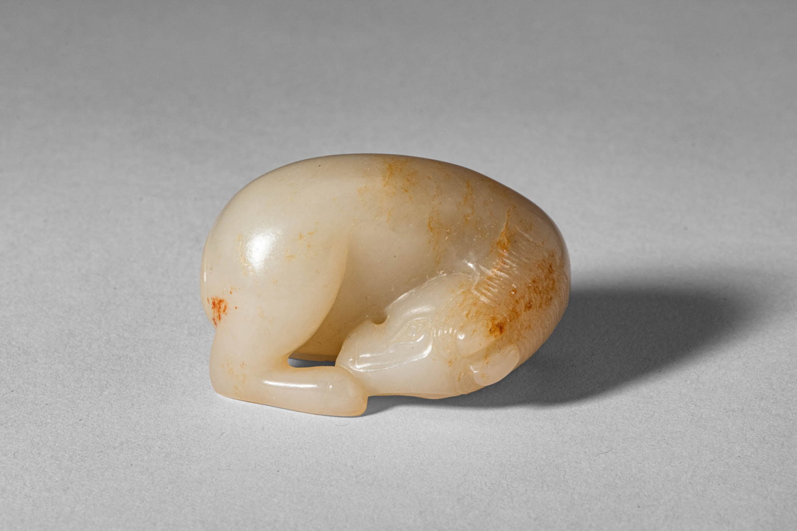 CHINESE WHITE JADE OF RECUMBENT HORSE: Mid Qing Dynasty, Ca. 18th century AD. >A carved jade figure of a recumbent horse, depicted in a naturalistic curled pose with legs tucked beneath the body and head turned gently to one side. The form