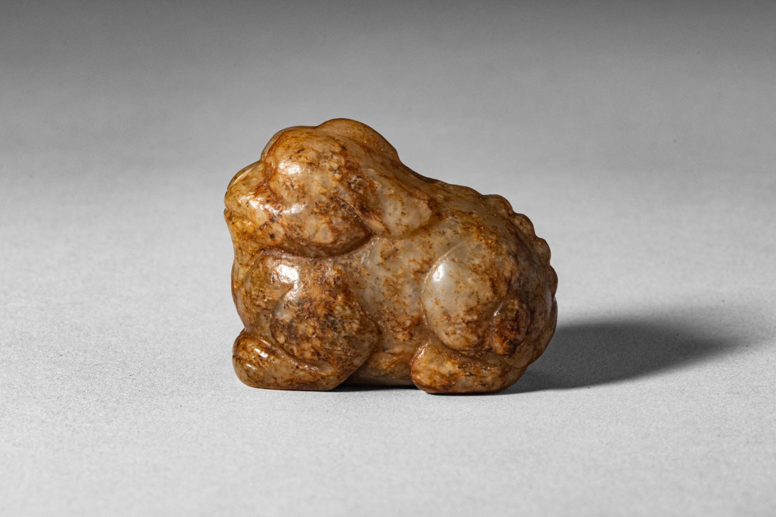 CHINESE WHITE JADE WITH RUSSET OF RECUMBENT BEAST: Ming Dynasty, Ca. AD 1368 - 1644, or earlier. >A compact jade carving depicting a recumbent mythical beast, shown with legs tucked beneath its body and head slightly raised in a composed, watchful pos