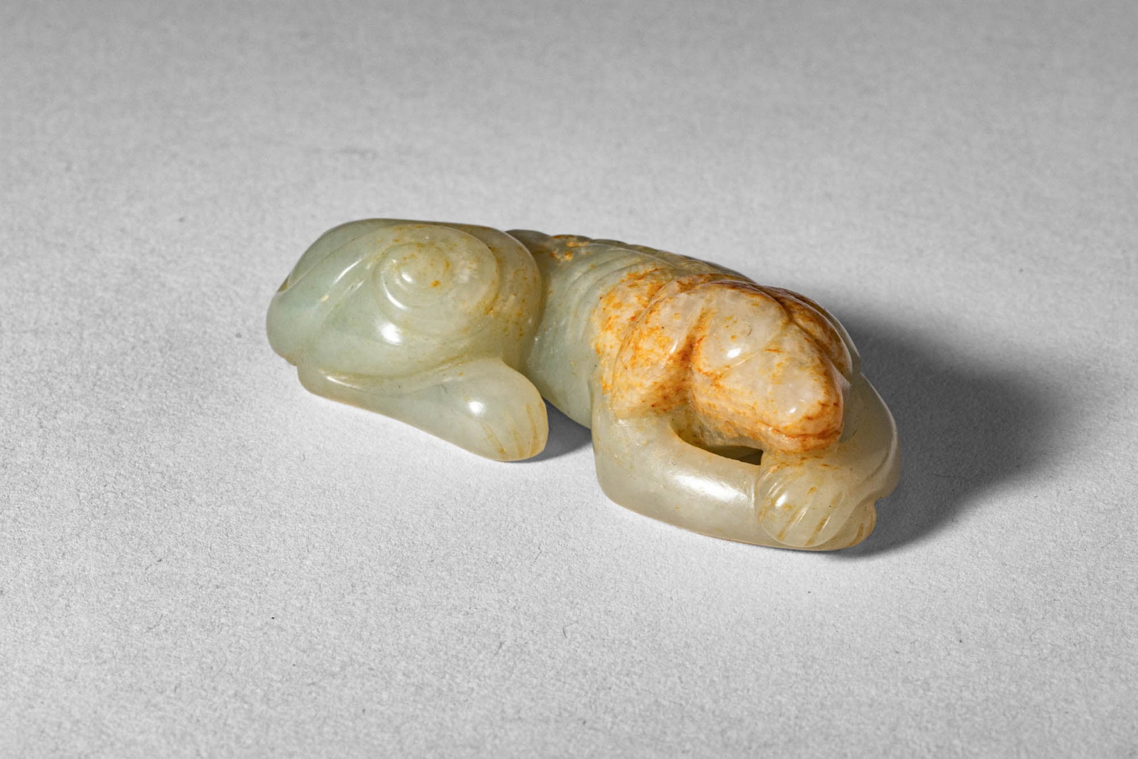 CHINESE CELADON JADE HOUND: Ca. 19th century AD. >A finely carved jade figure of a recumbent hound, shown lying with its body stretched forward and head resting low in a relaxed pose. The form is compact and smoothly modelled, w