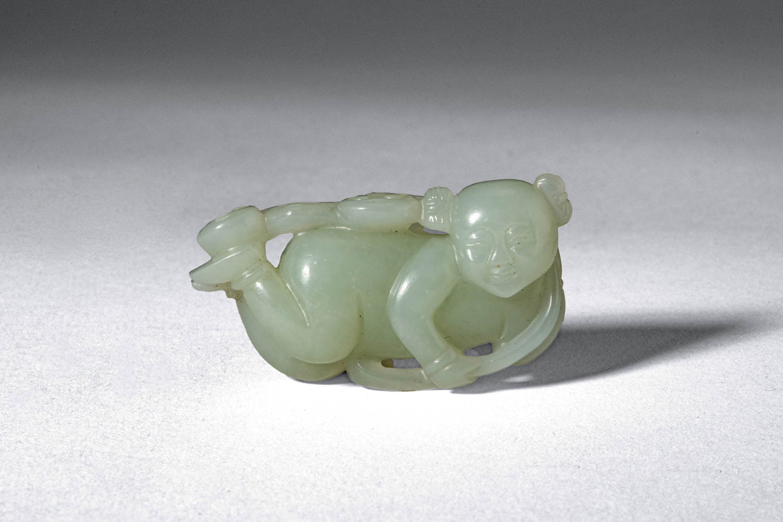 CHINESE PALE CELADON JADE BOY: Ca. 19th century AD. >A carved jade figure depicting a playful boy reclining in a relaxed pose, his body rendered with rounded, naturalistic modelling. The child is shown with one leg raised and arms 