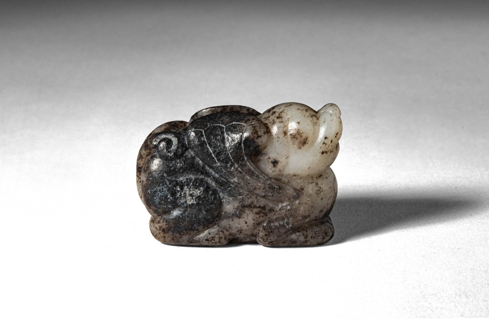 CHINESE WHITE JADE OF RECUMBENT WINGED BEAST: Ming Dynasty, Ca. AD 1368 - 1644, or earlier. >A carved jade figure depicting a recumbent mythical beast, the compact body rendered with legs tucked beneath and head raised in an upward poised attitud