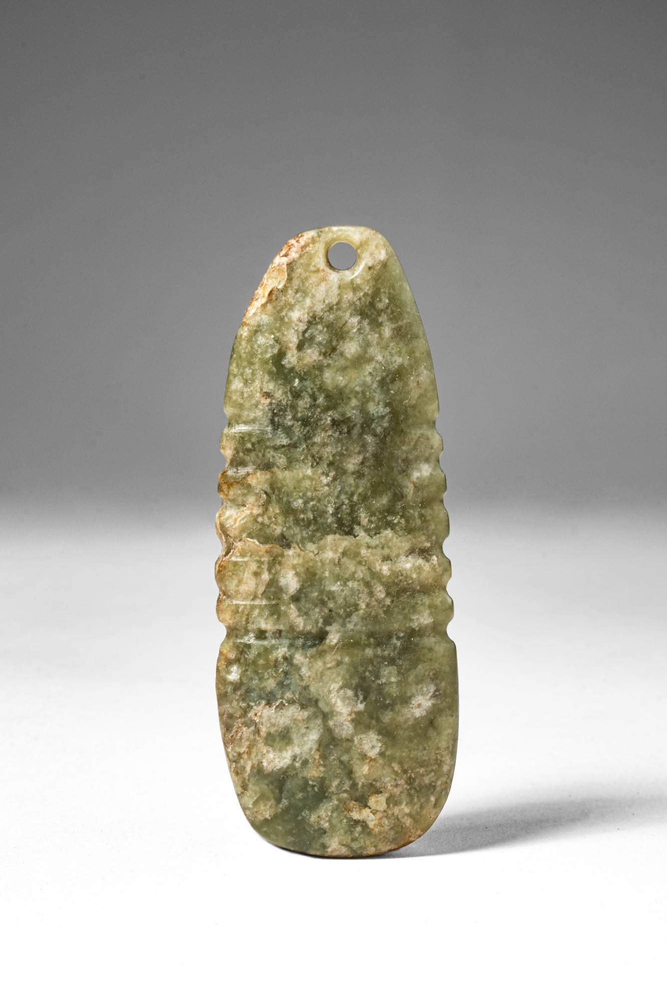 CHINESE JADE PENDANT: . >A jade pendant of elongated oval form with a rounded terminal, pierced at the top for suspension, the stone carved with subtle raised bands across the body. The surface is worked smooth, the pale g